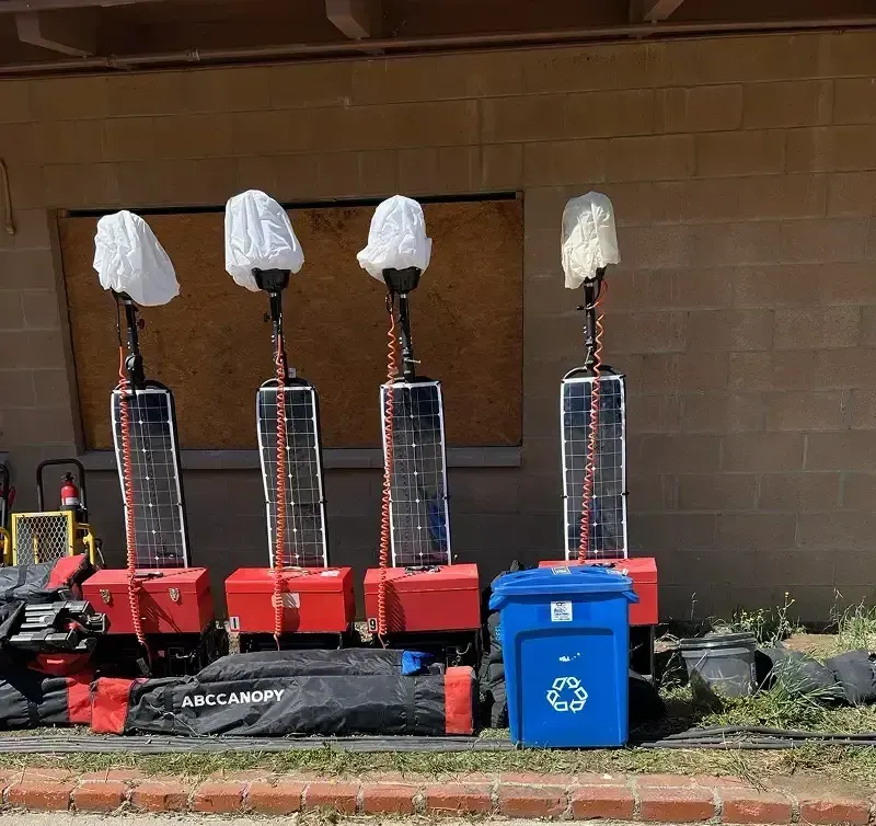 Four solar-powered light towers with white covers, next to a blue recycling bin and equipment in front of a brick building.