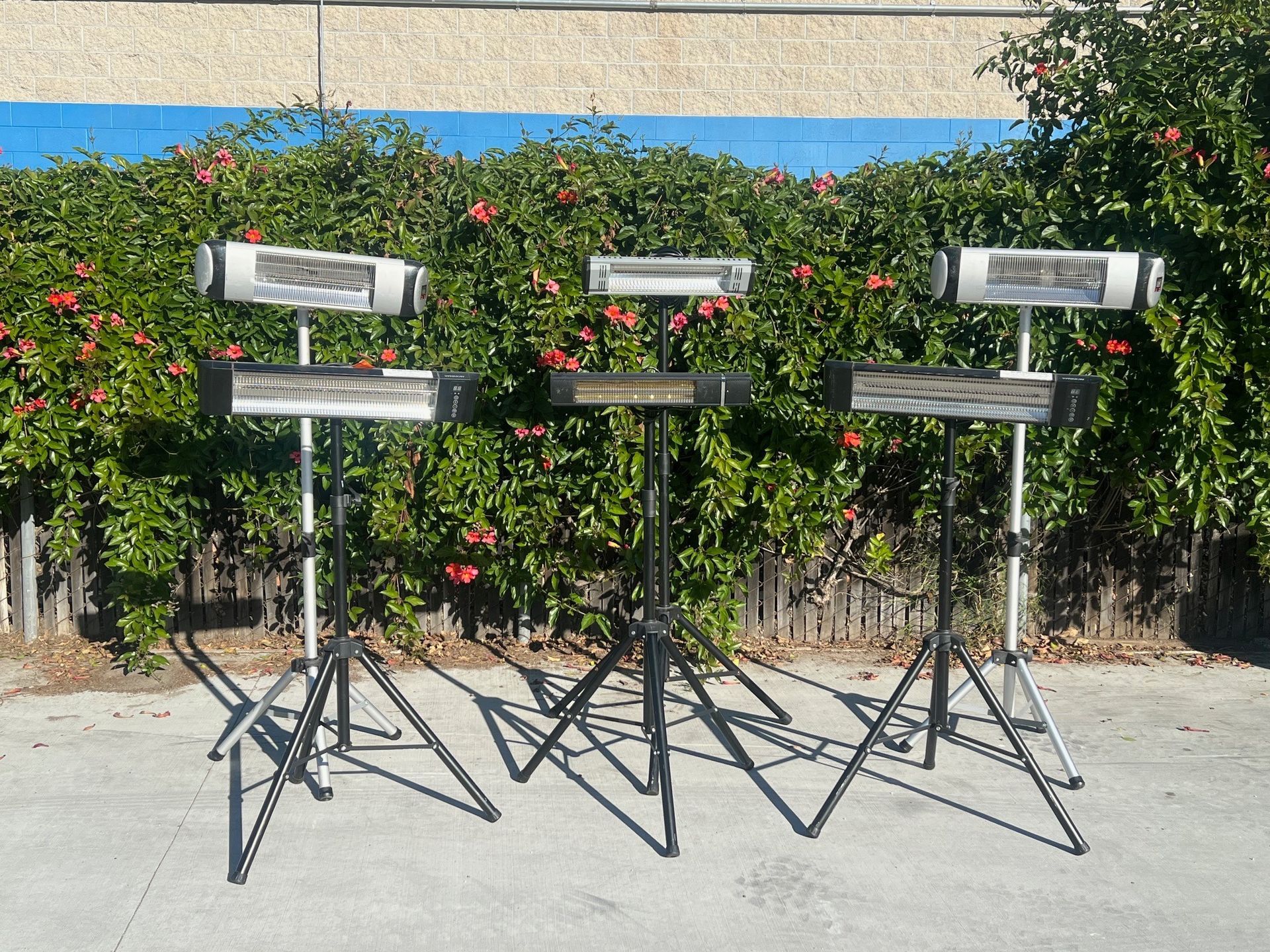 Three outdoor heaters on stands, in front of a hedge and blue wall.