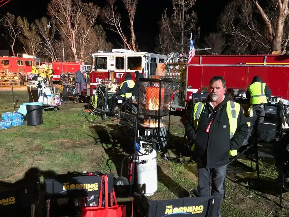 A man in a black jacket stands in front of several fire trucks at night, with crew members and equipment in the background.