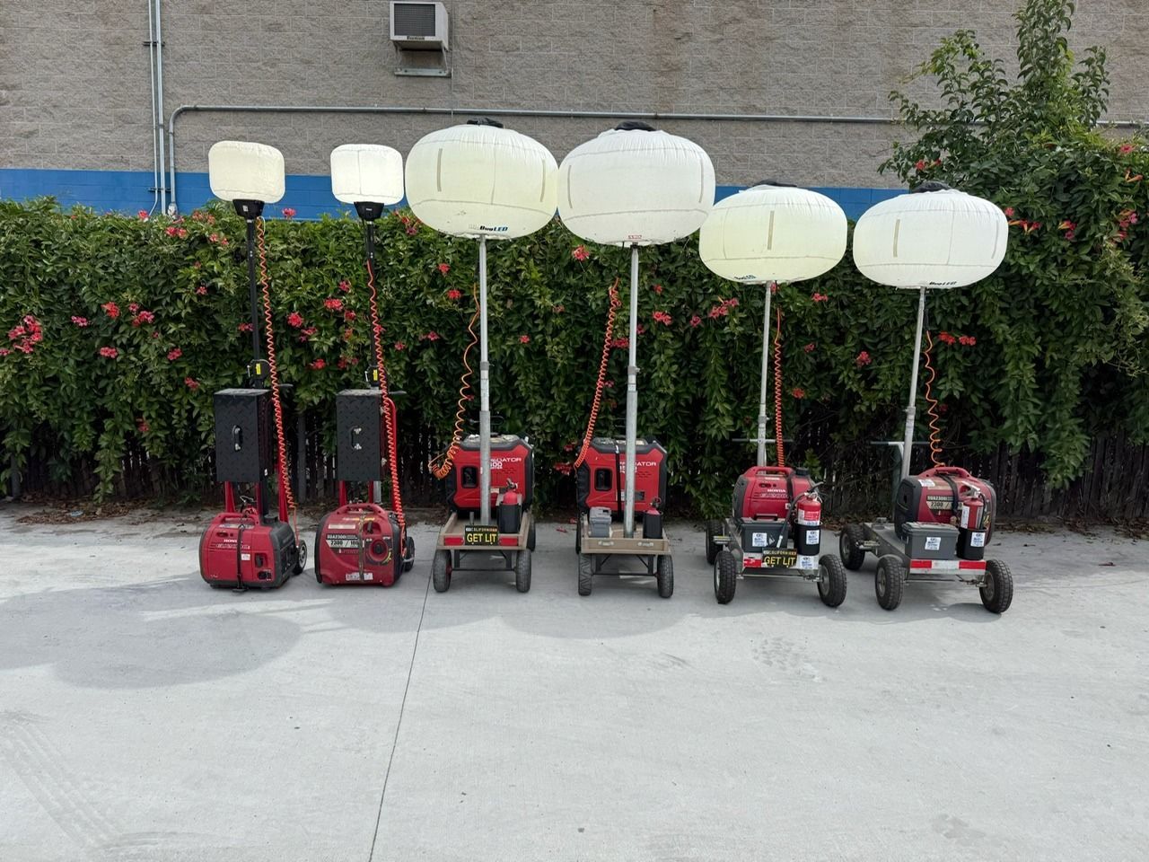 Row of portable lighting units with red bases, white balloon-shaped lights, and metal poles. They are outside next to greenery.