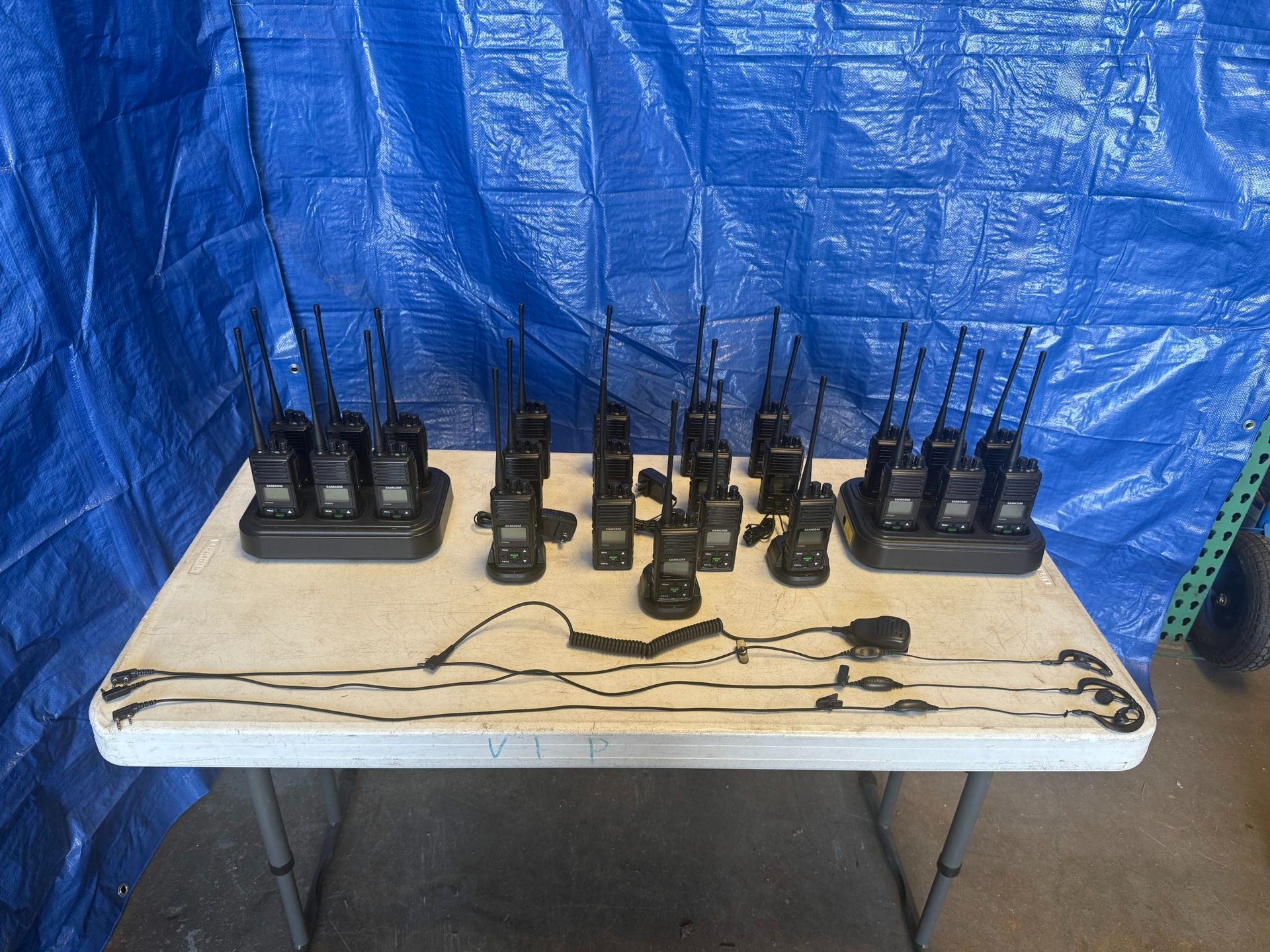 Black walkie-talkies on a white table. Some are in chargers. Blue tarp background.