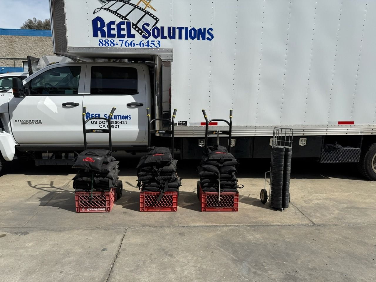 A white moving truck with “Reel Solutions” logo has three hand trucks with black bags and rolled equipment.