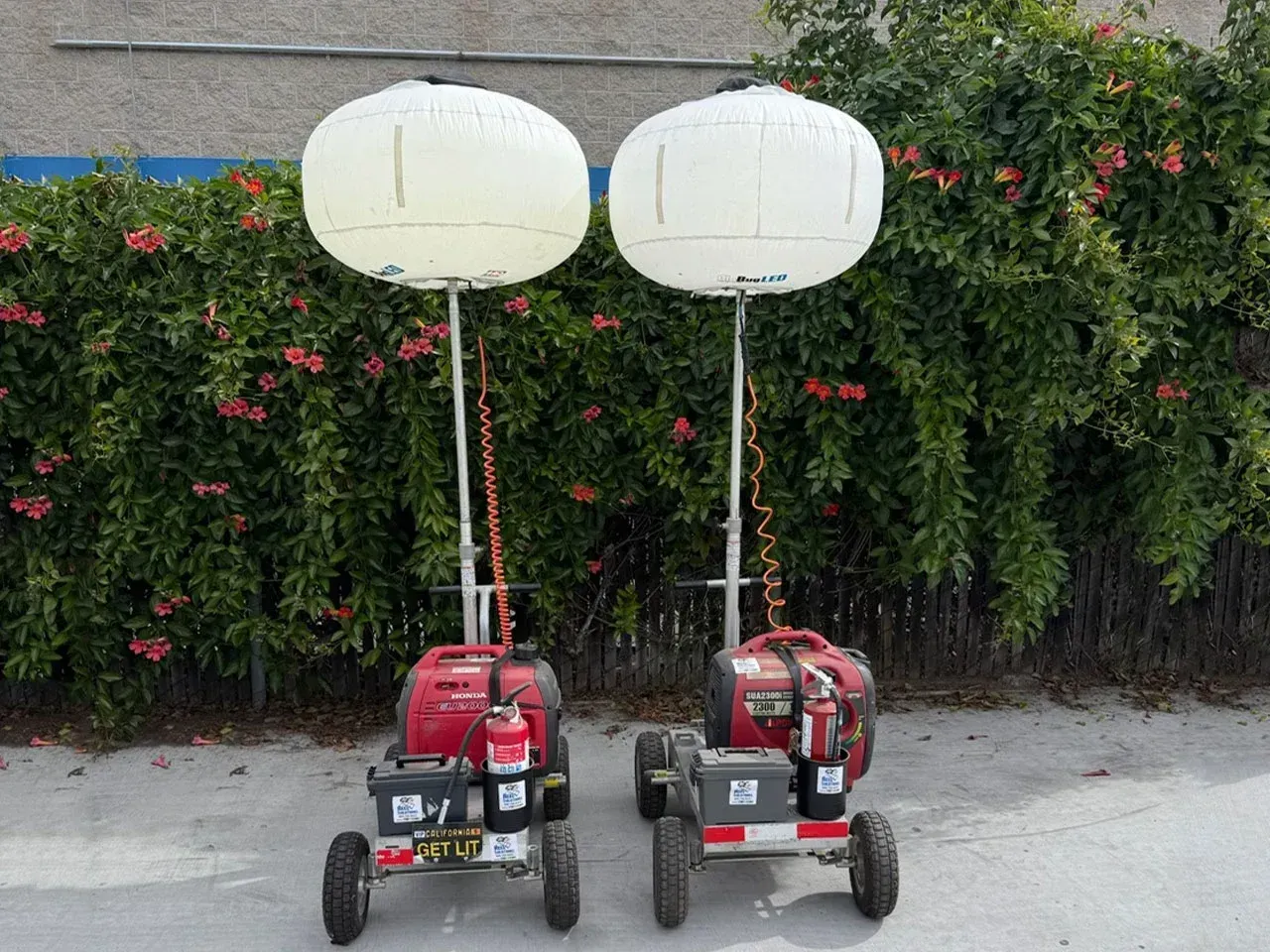 Two portable, red-and-gray generators with tall poles topped by white, round lights. Parked in front of bushes.