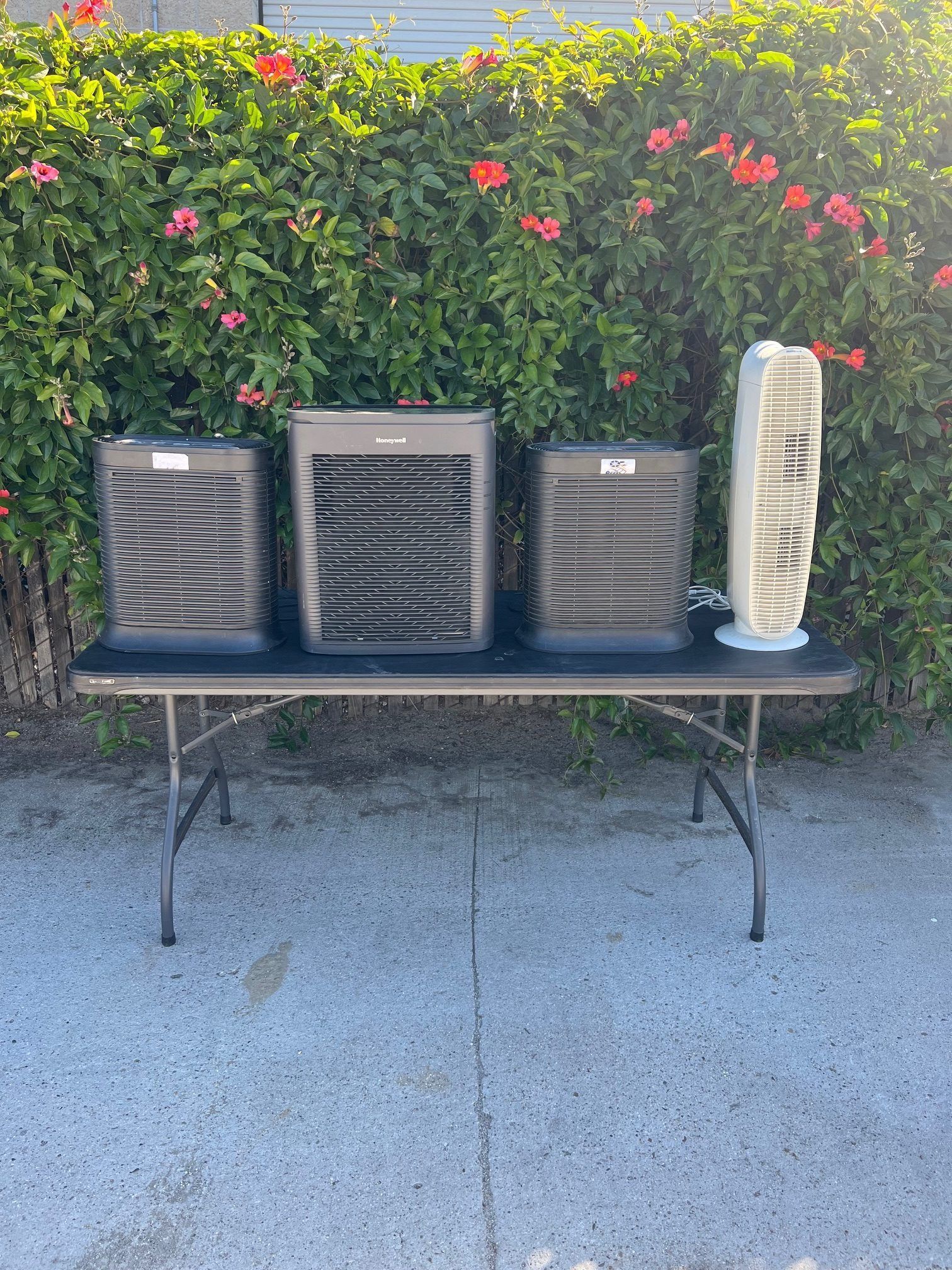 Four air purifiers and a fan on a black table against a backdrop of green foliage with red flowers.