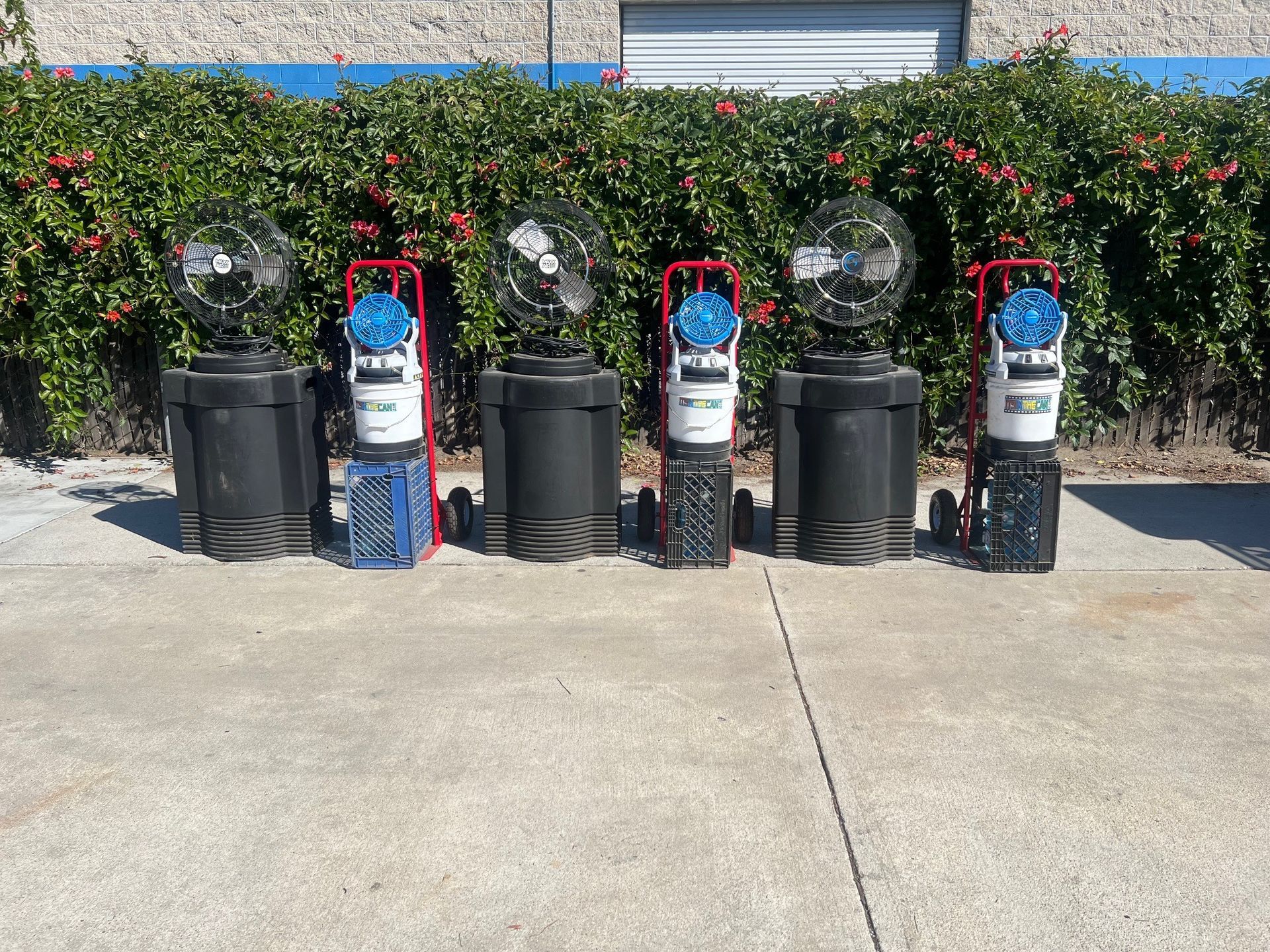 Five portable misting fans lined up in front of a green hedge and building.