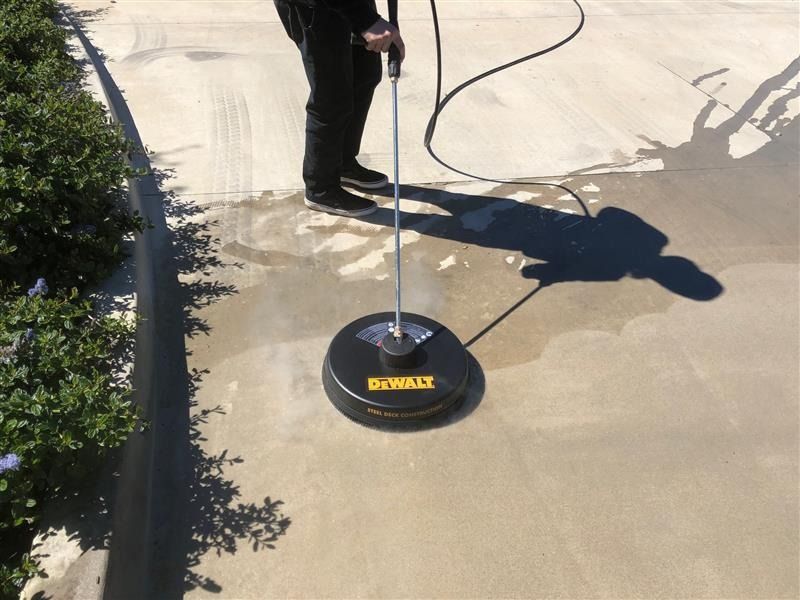 Person using a pressure washer with circular attachment to clean a concrete surface outdoors.