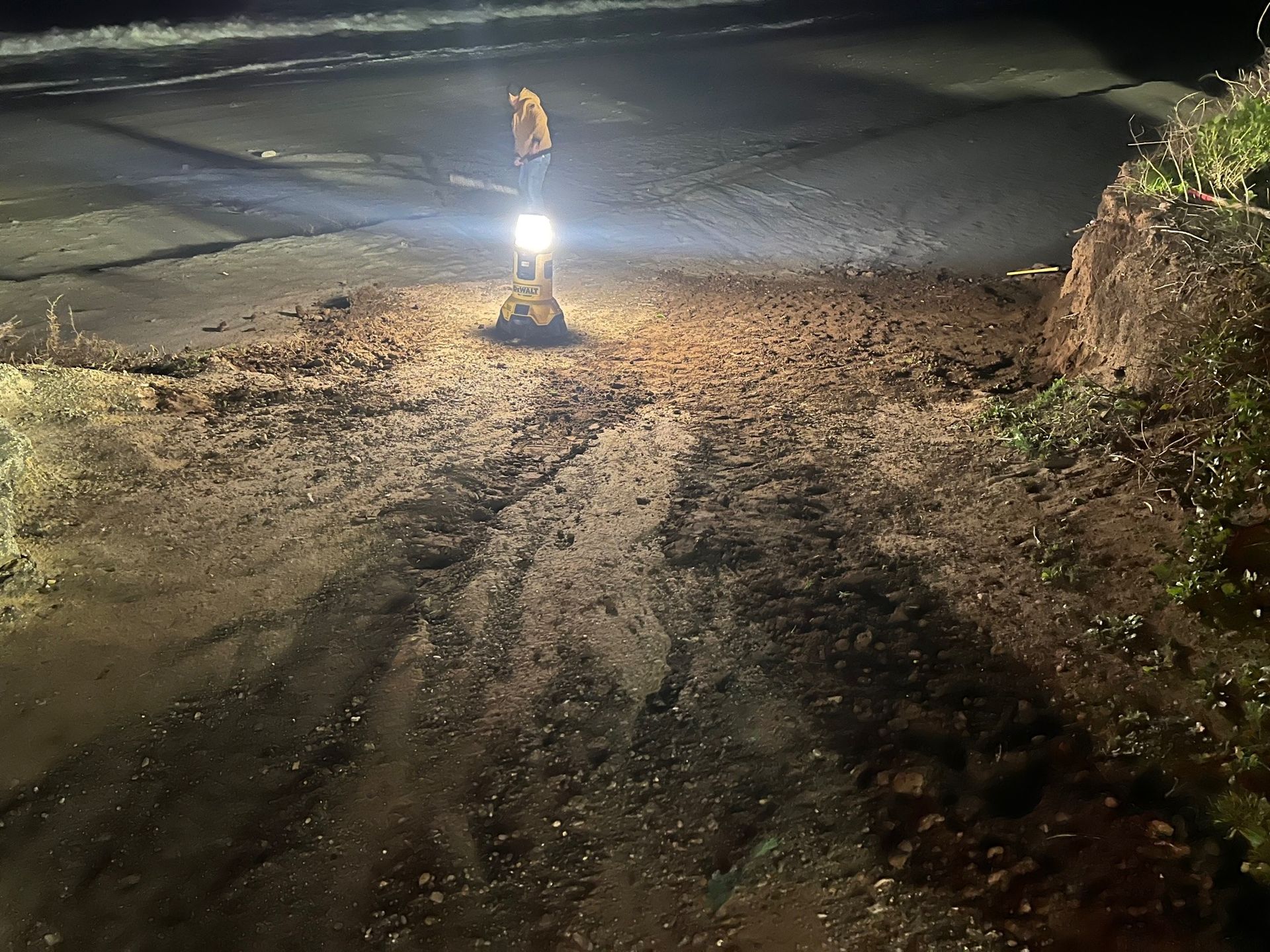 A construction worker uses a light in the dark to examine a damaged road covered in loose dirt.