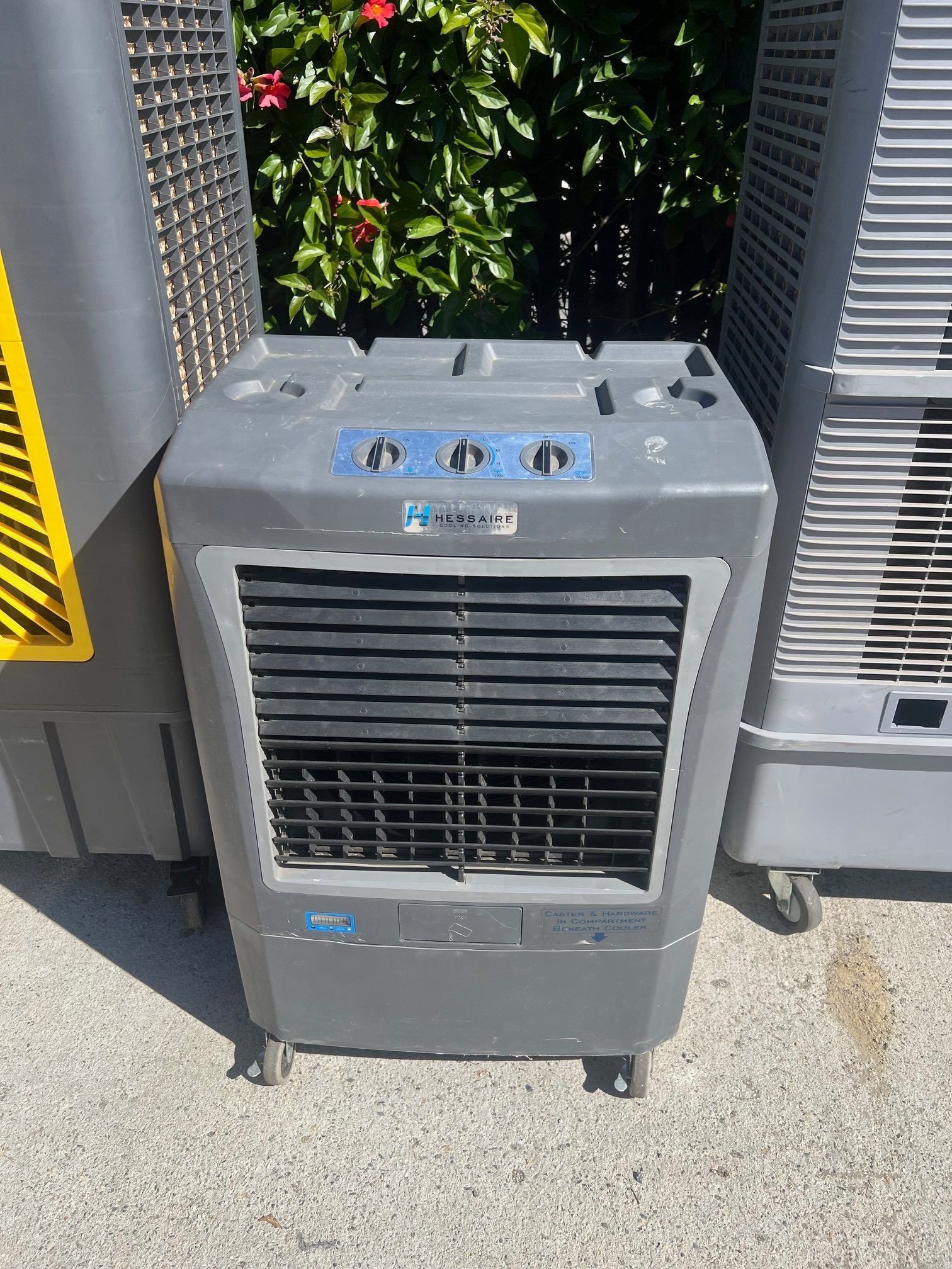 Gray evaporative cooler with knobs, dark front vent, and wheels, outdoors.