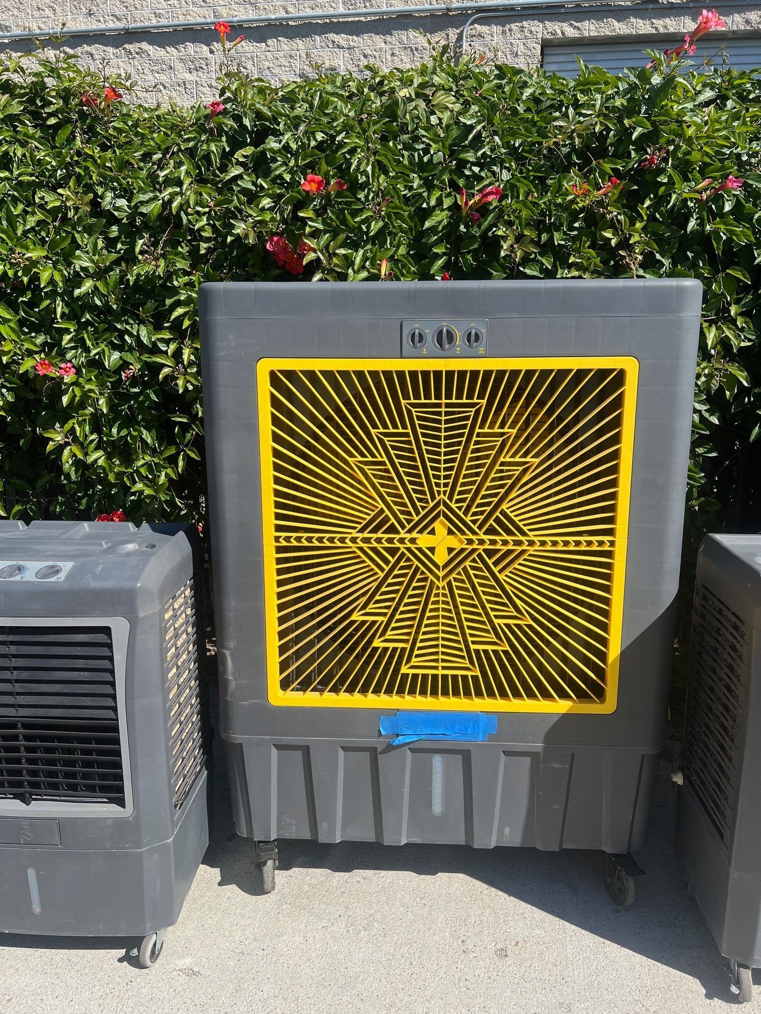 Three gray evaporative coolers on wheels in front of a green hedge; center one has a yellow and black geometric design.