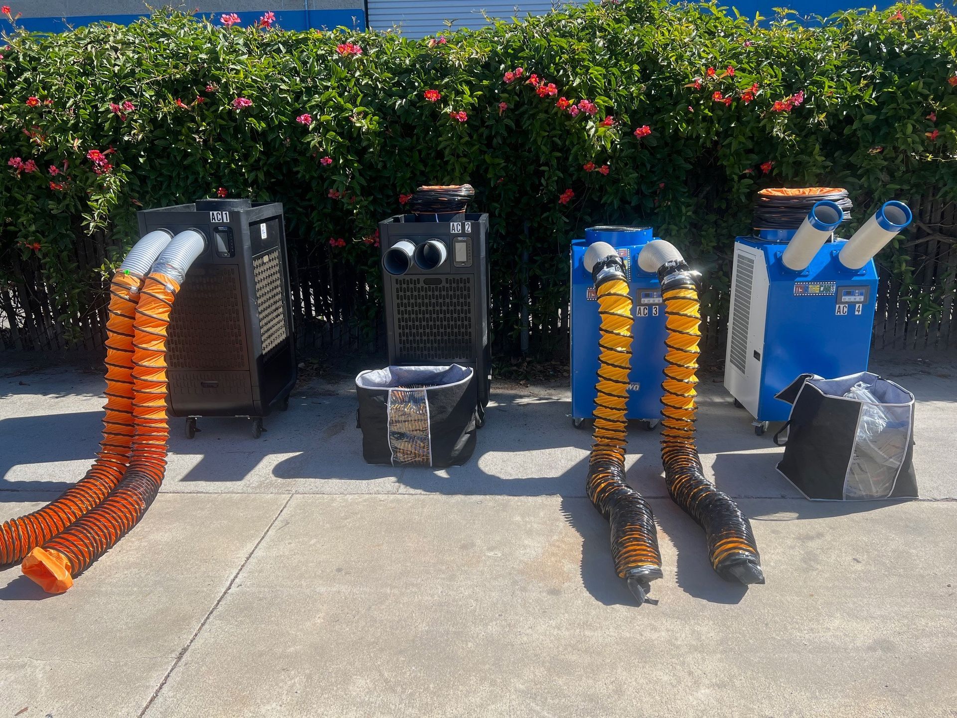 Four industrial air conditioning units with large orange and yellow hoses outdoors near greenery.