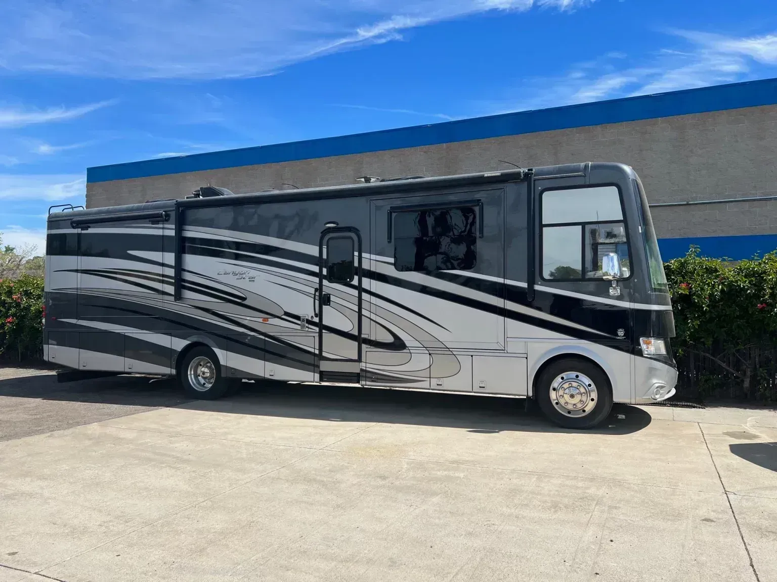Gray and white recreational vehicle parked on concrete, against a blue and gray building under a sunny sky.