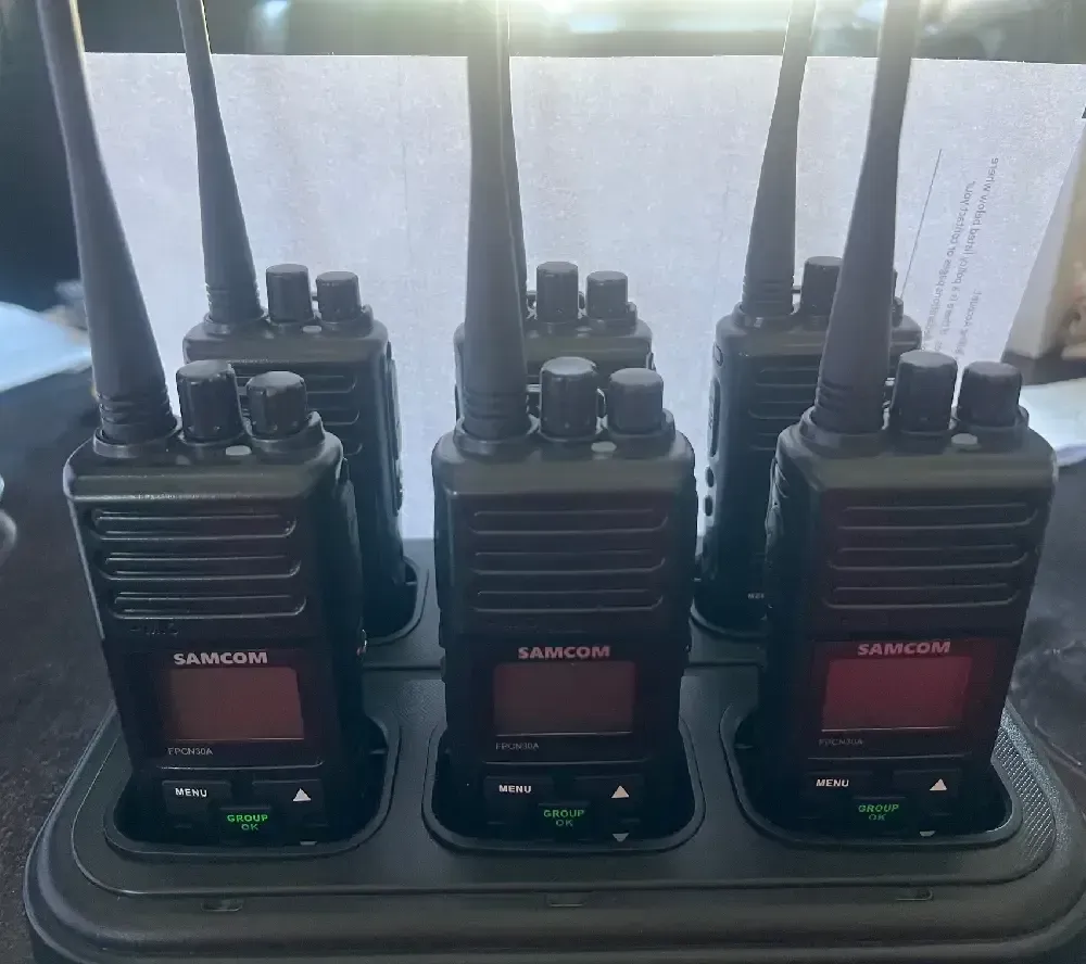 Six black walkie-talkies in a charging station, each with an antenna and screen, displaying 