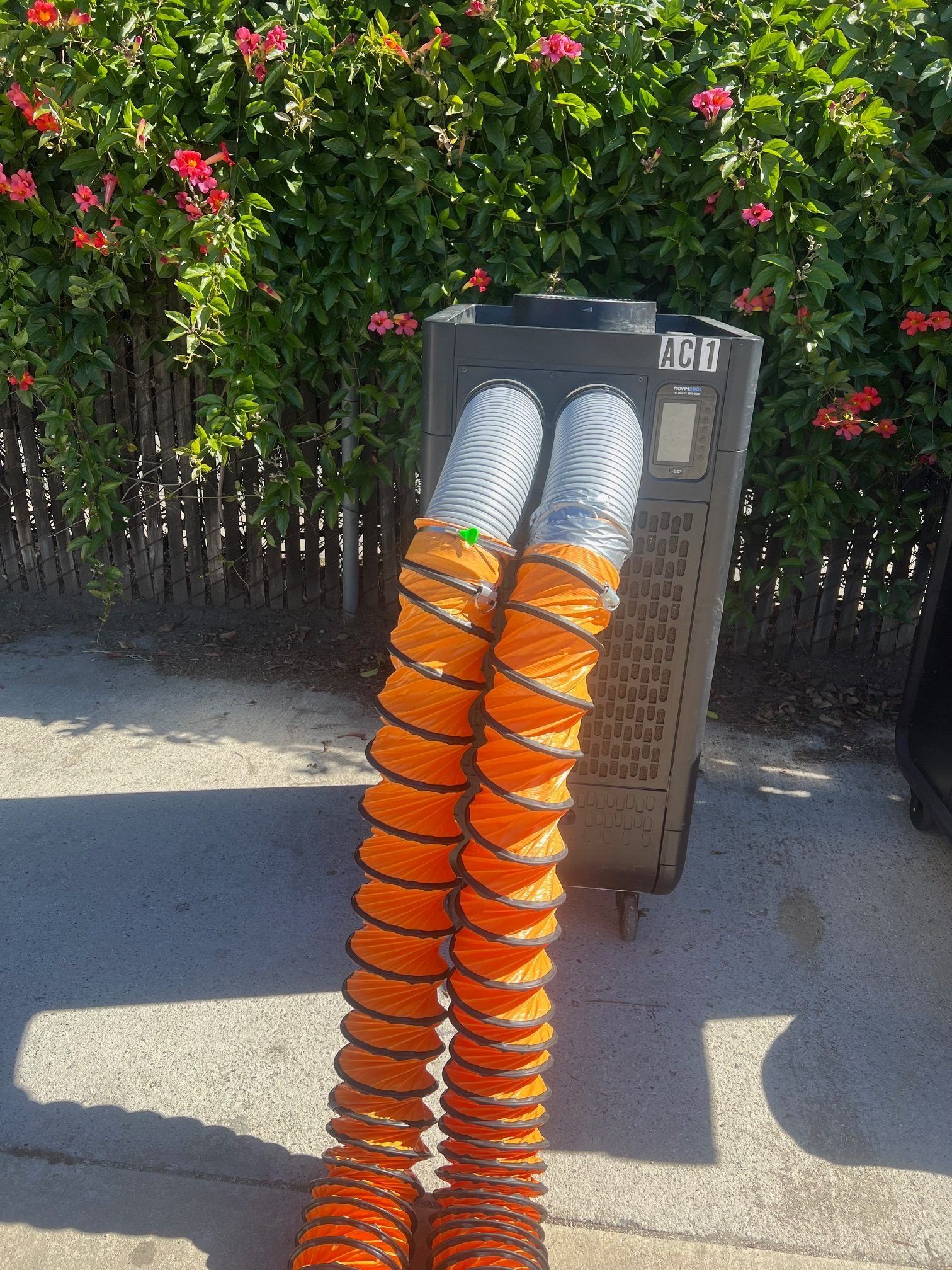 Two orange flexible tubes connected to a dark gray machine, with flowers and a fence in the background.
