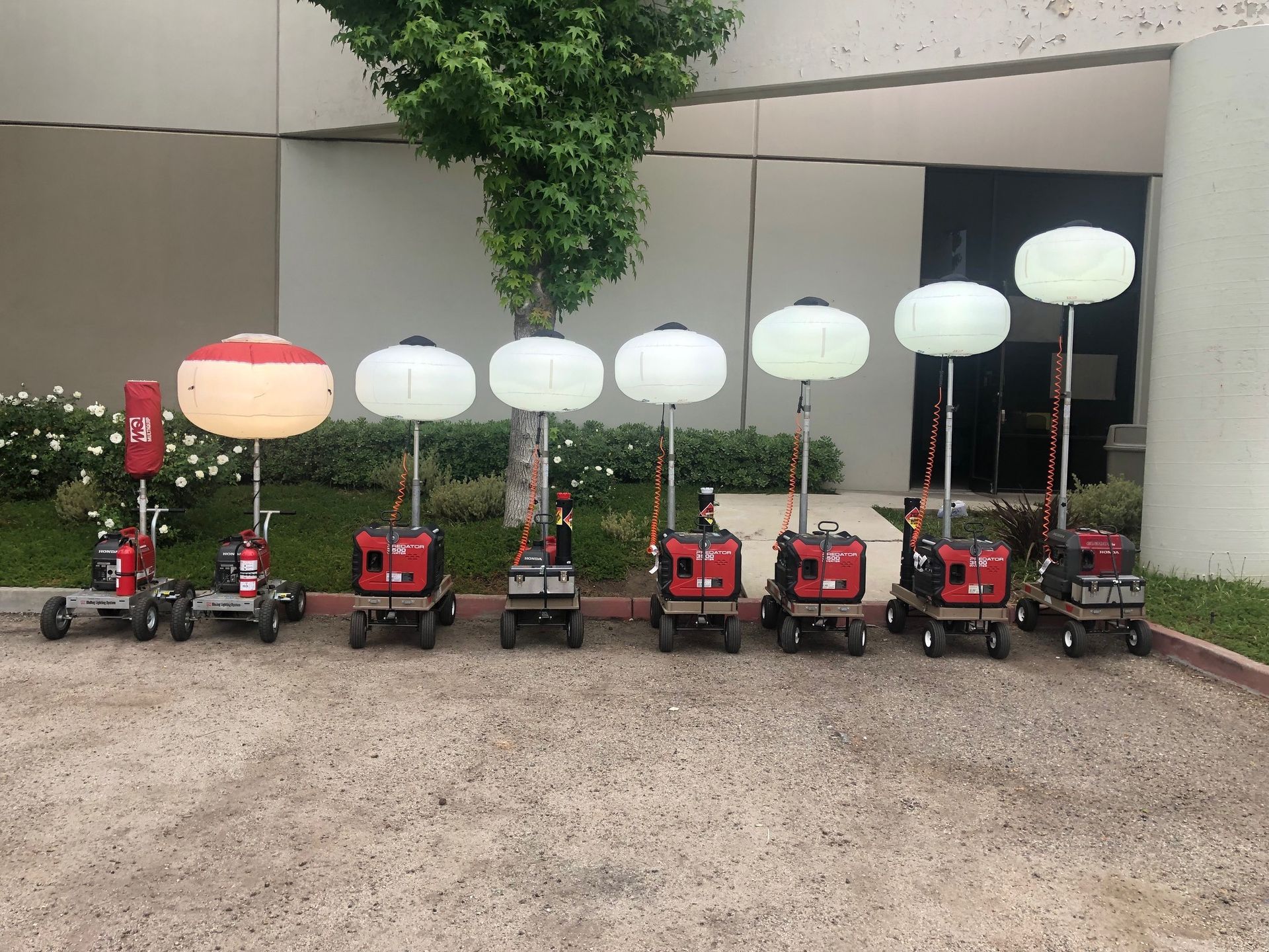 Line of red portable light towers with white or orange globes, parked on asphalt.