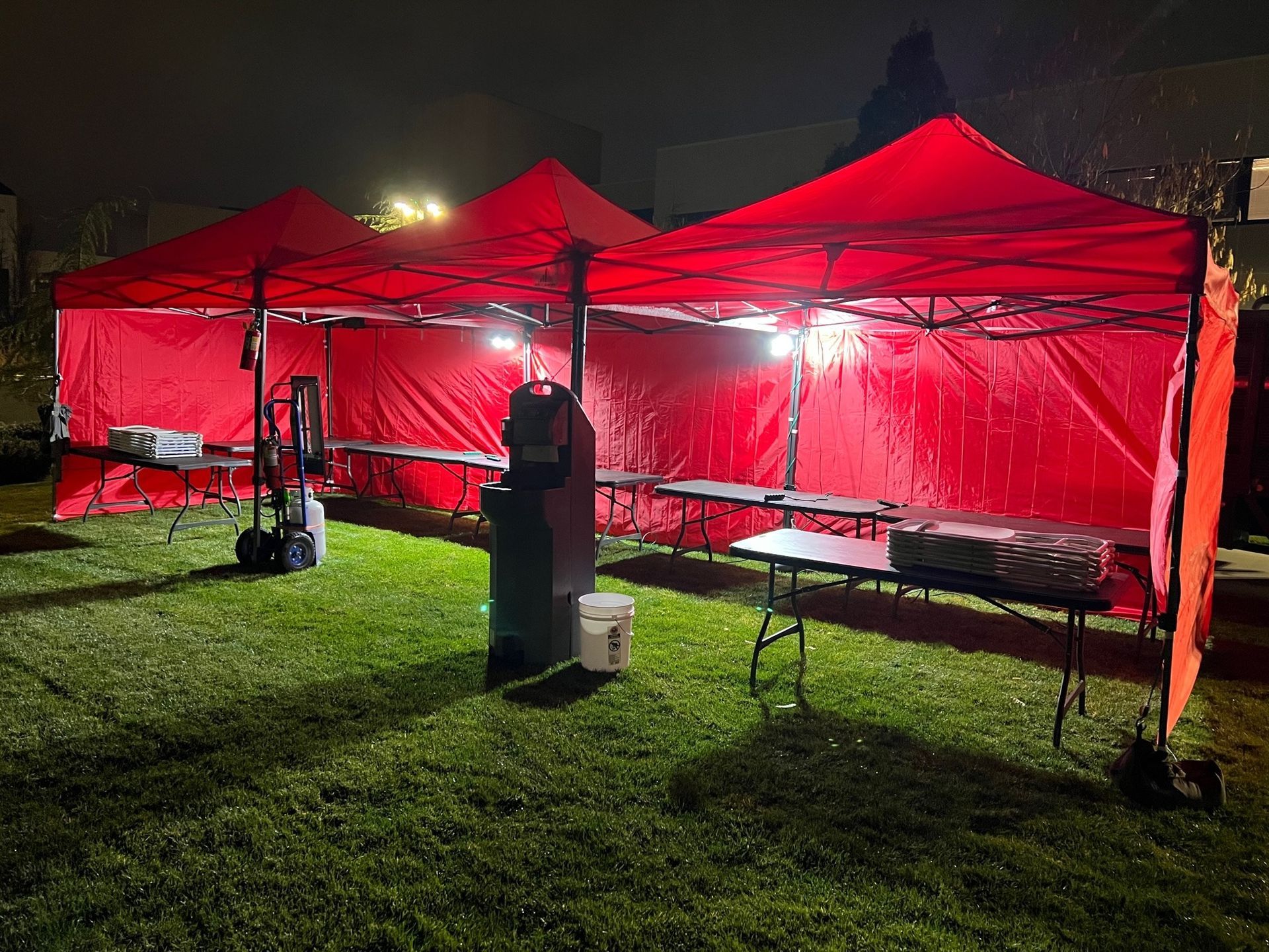 Three illuminated red canopies set up on grass at night, with tables and equipment inside.