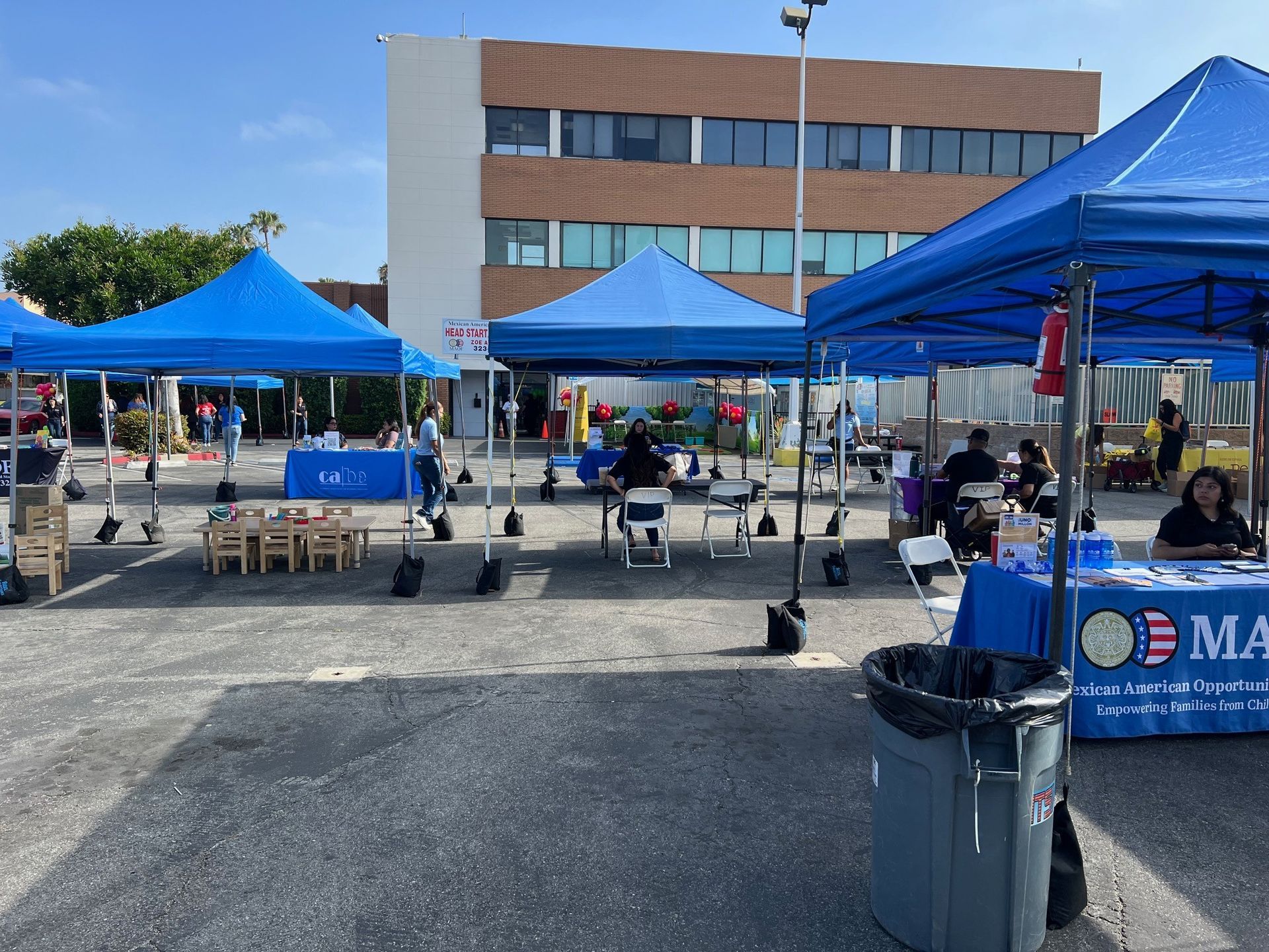 Blue canopy tents set up outdoors for an event with people. A multi-story building is in the background.