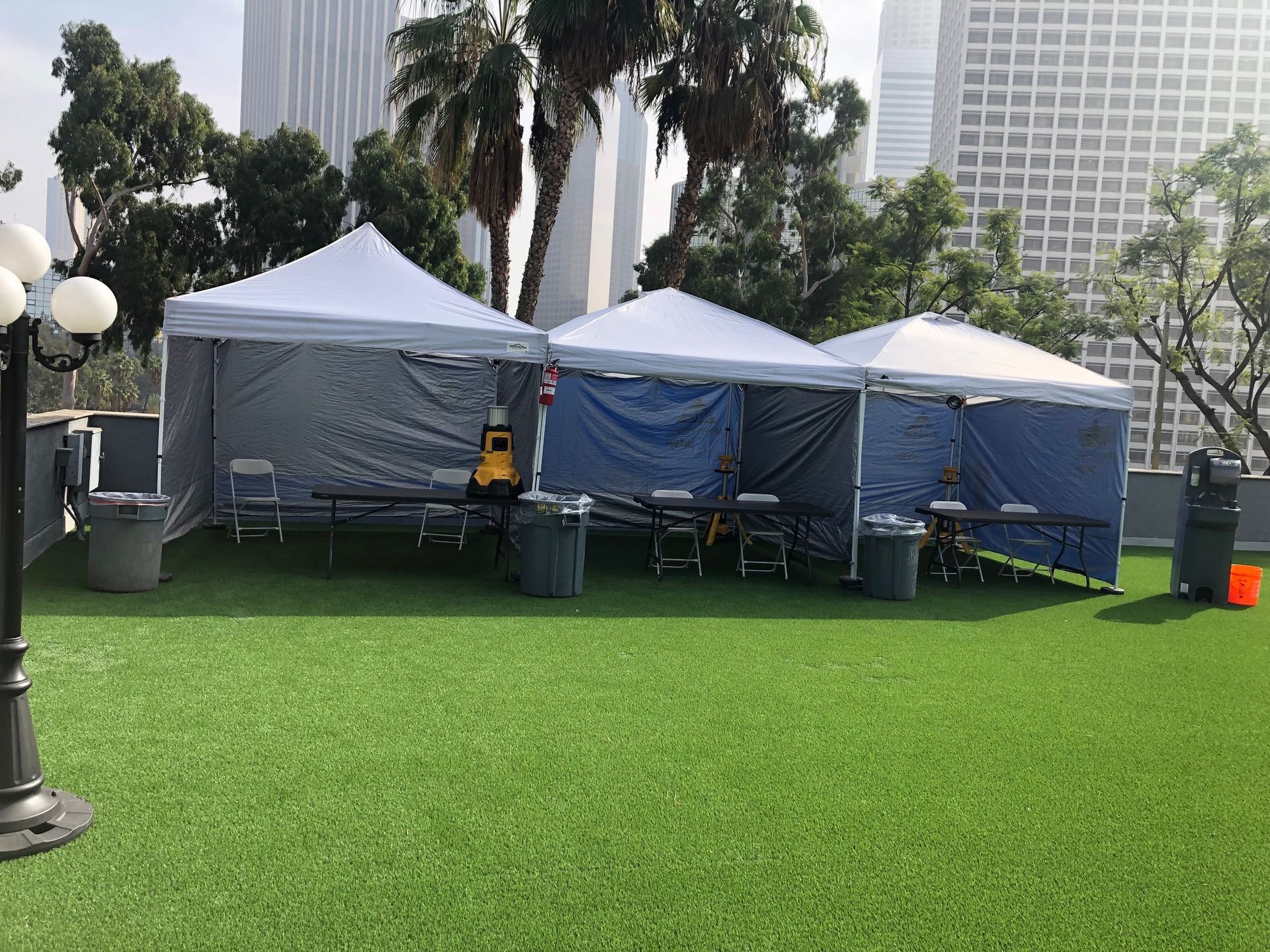 Three white tents set up on artificial grass, tables, chairs, and trash cans. City buildings and palm trees in background.
