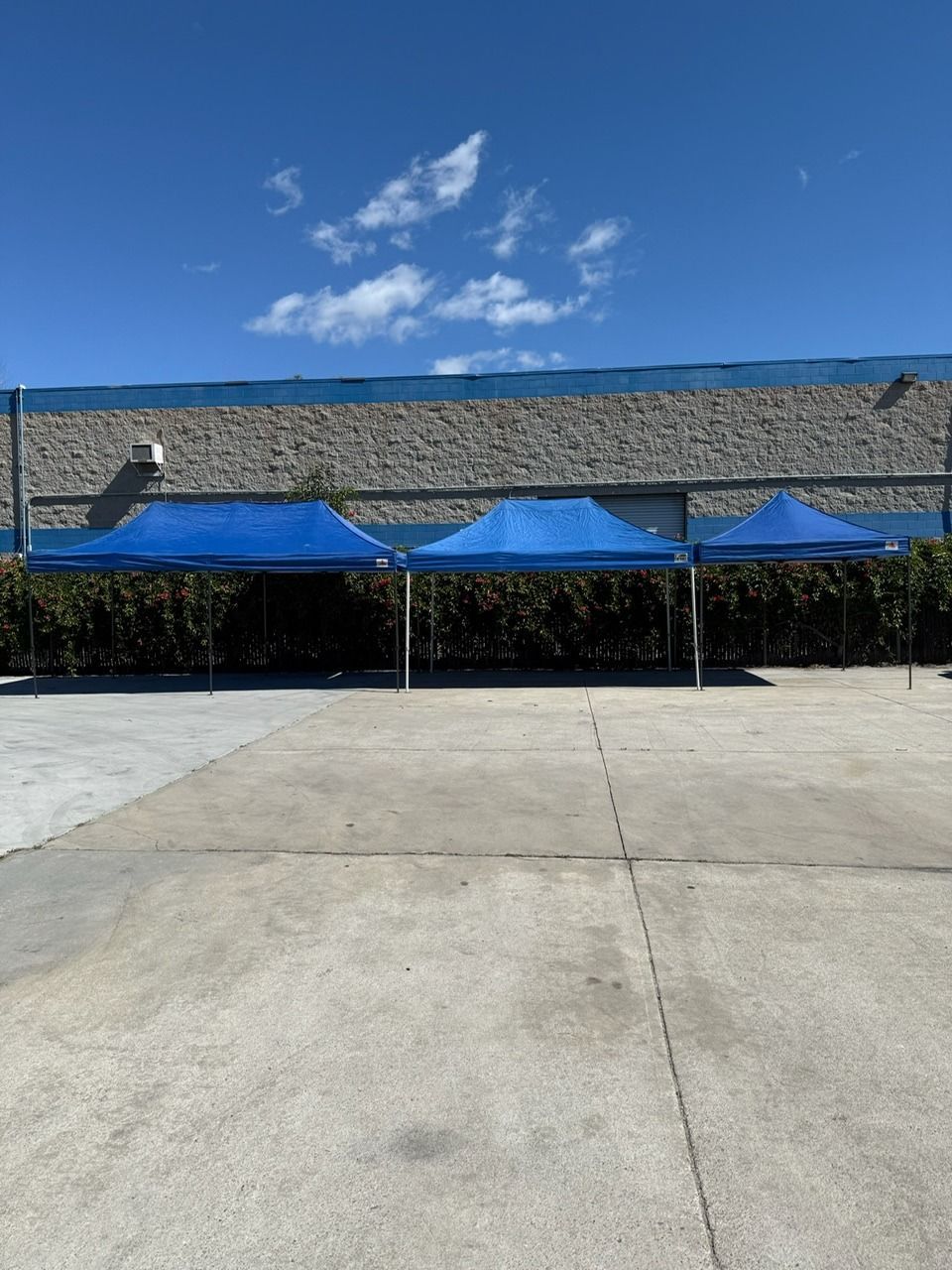 Three blue pop-up tents lined up on a concrete surface, against a brick building under a blue sky.
