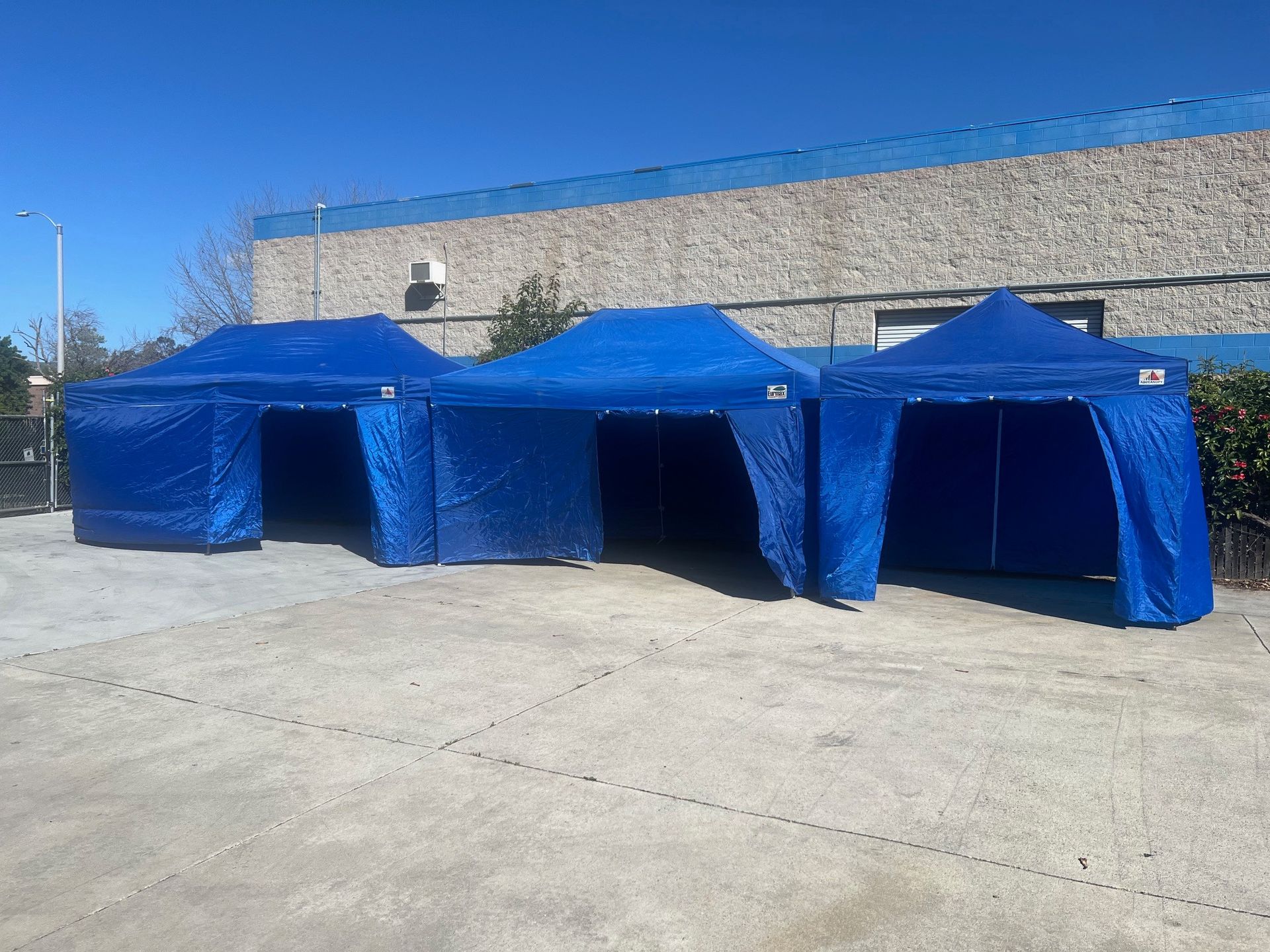 Three blue pop-up tents lined up outside a building on a sunny day.