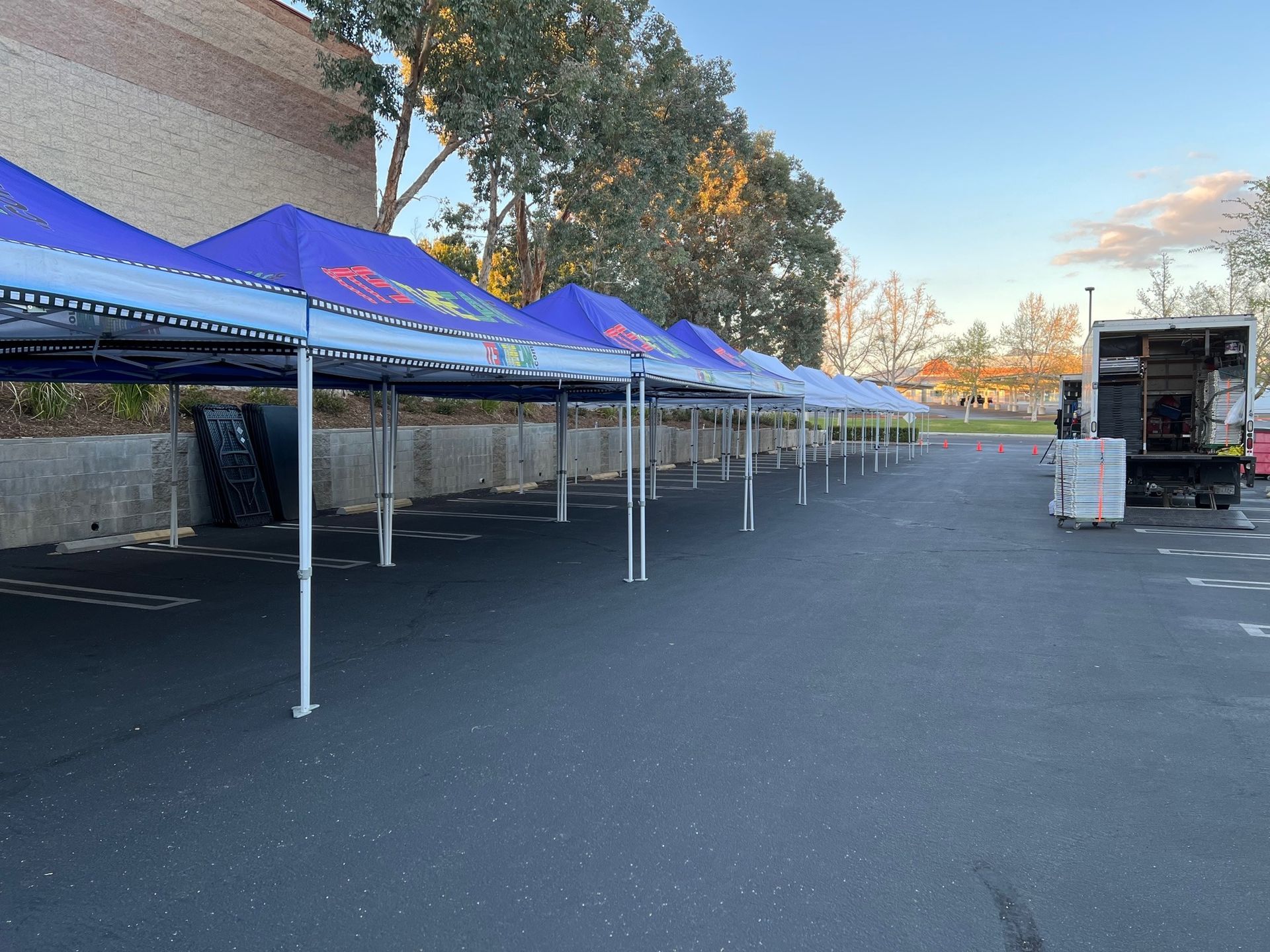 Row of blue canopy tents set up on asphalt, near a building and trees.
