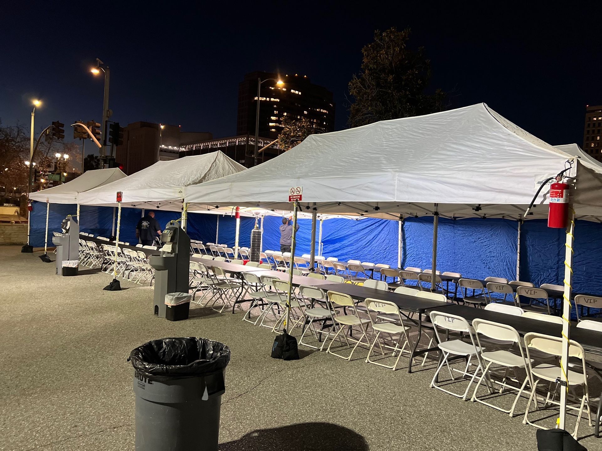 Row of white tents with tables and chairs set up outside. Blue backdrop, dark setting.