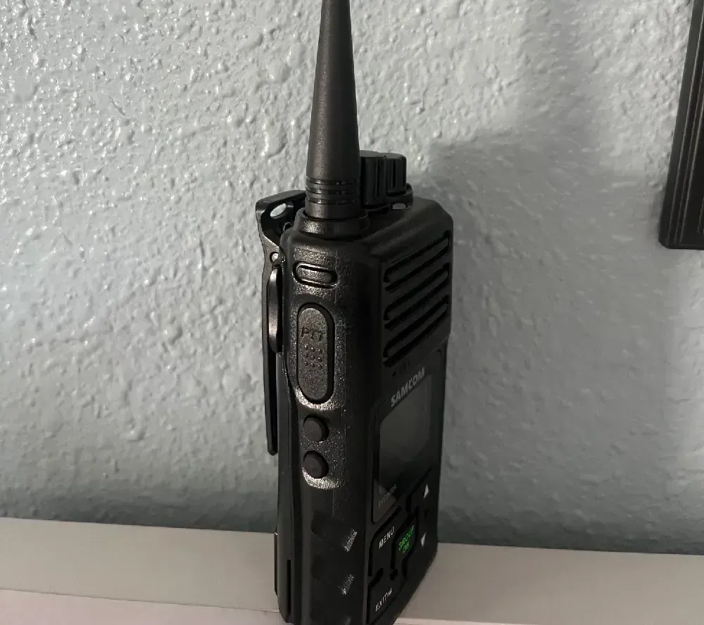 Black walkie-talkie with antenna and belt clip, against a textured blue wall.