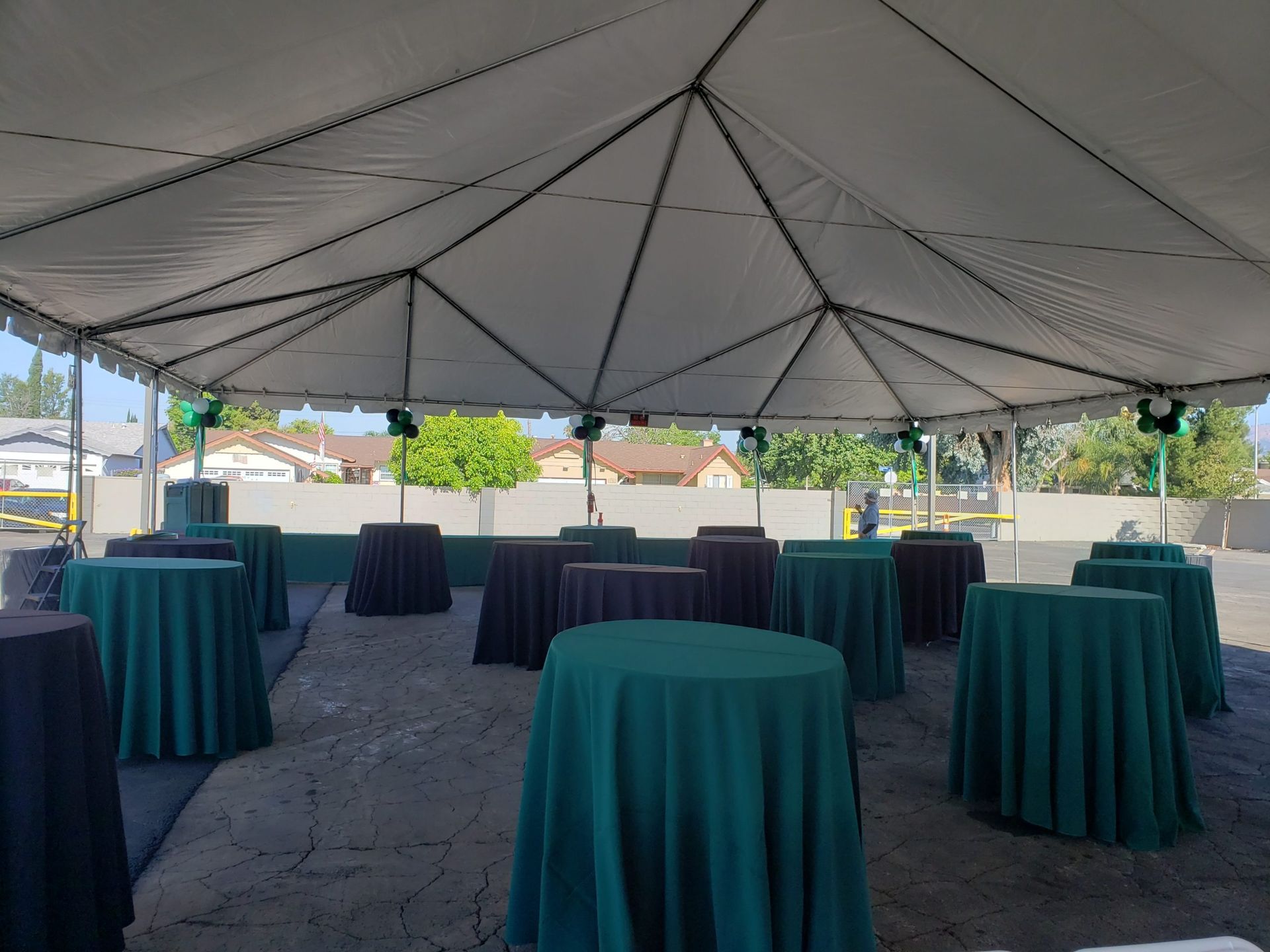 Tent-covered outdoor event with tables draped in green and brown cloth, set on a paved surface.