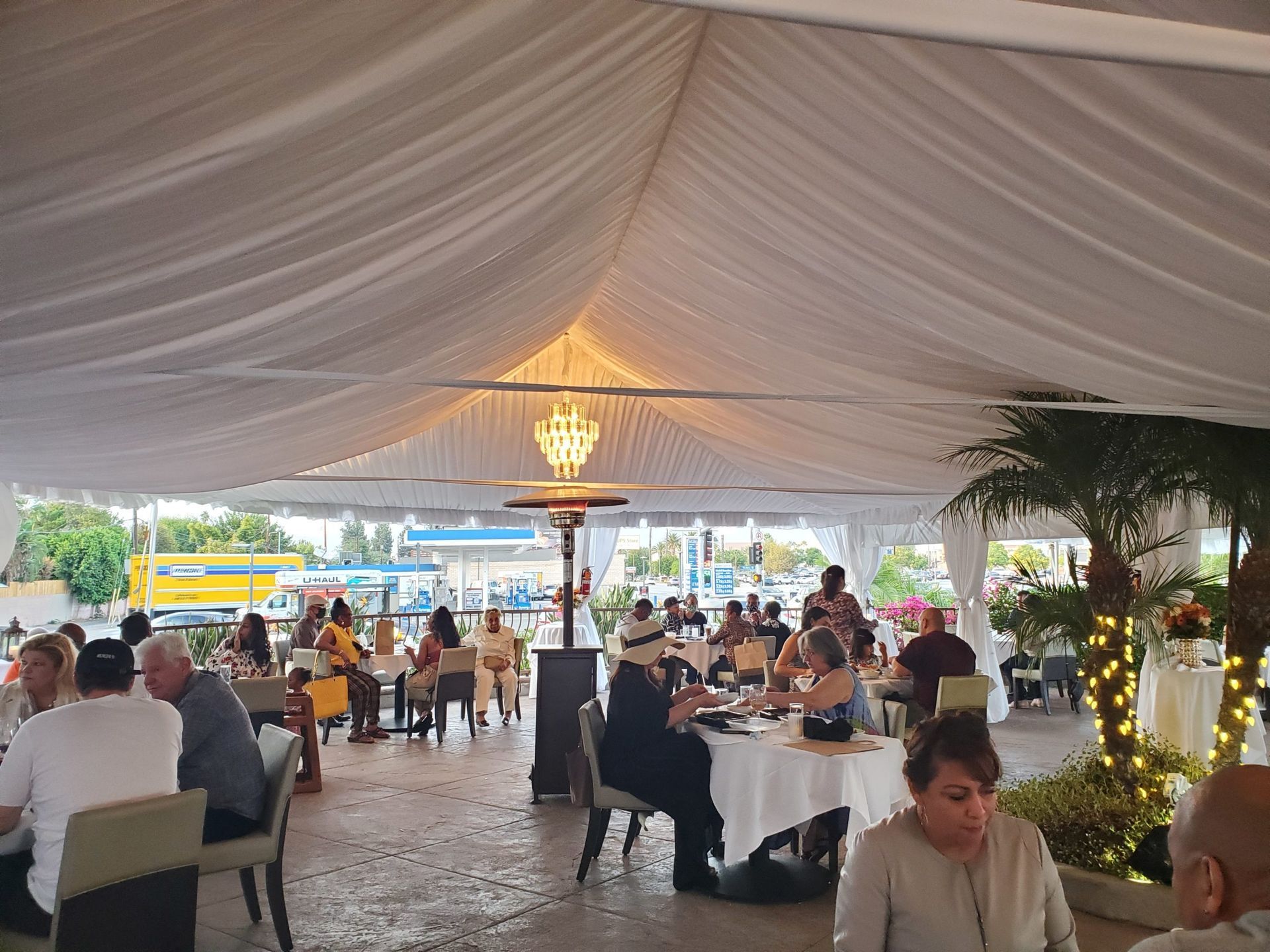 Outdoor restaurant scene under a white tent; people dining at tables, a chandelier hangs in the center.