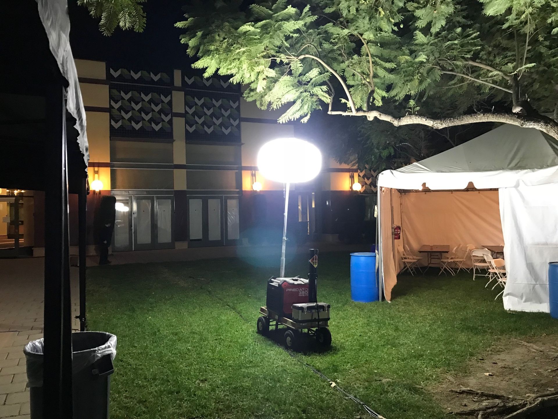 A bright, portable light illuminates a grassy area near a building and tent at night.