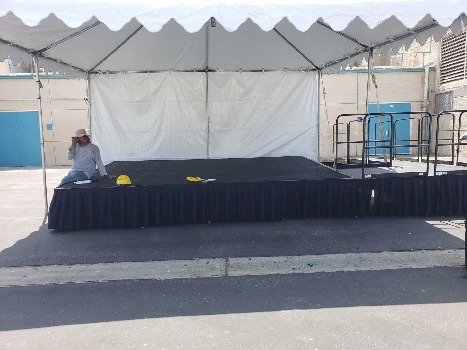 A man sitting at a table on a stage under a white tent. Black skirt covers the front of the stage.