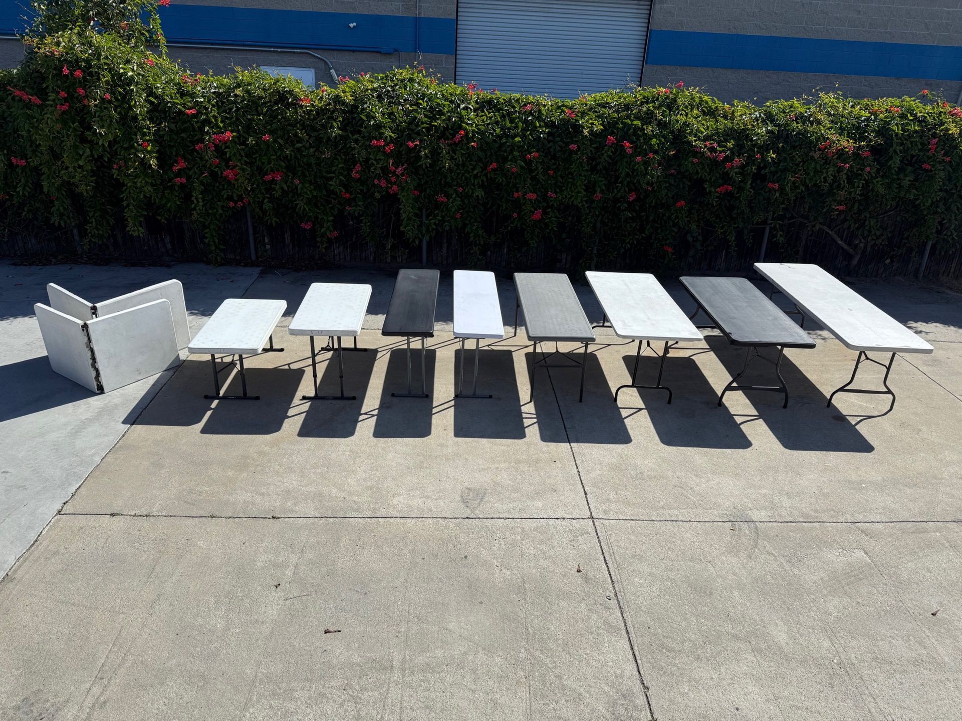 Tables of varying colors and sizes, lined up outdoors in front of a hedge.
