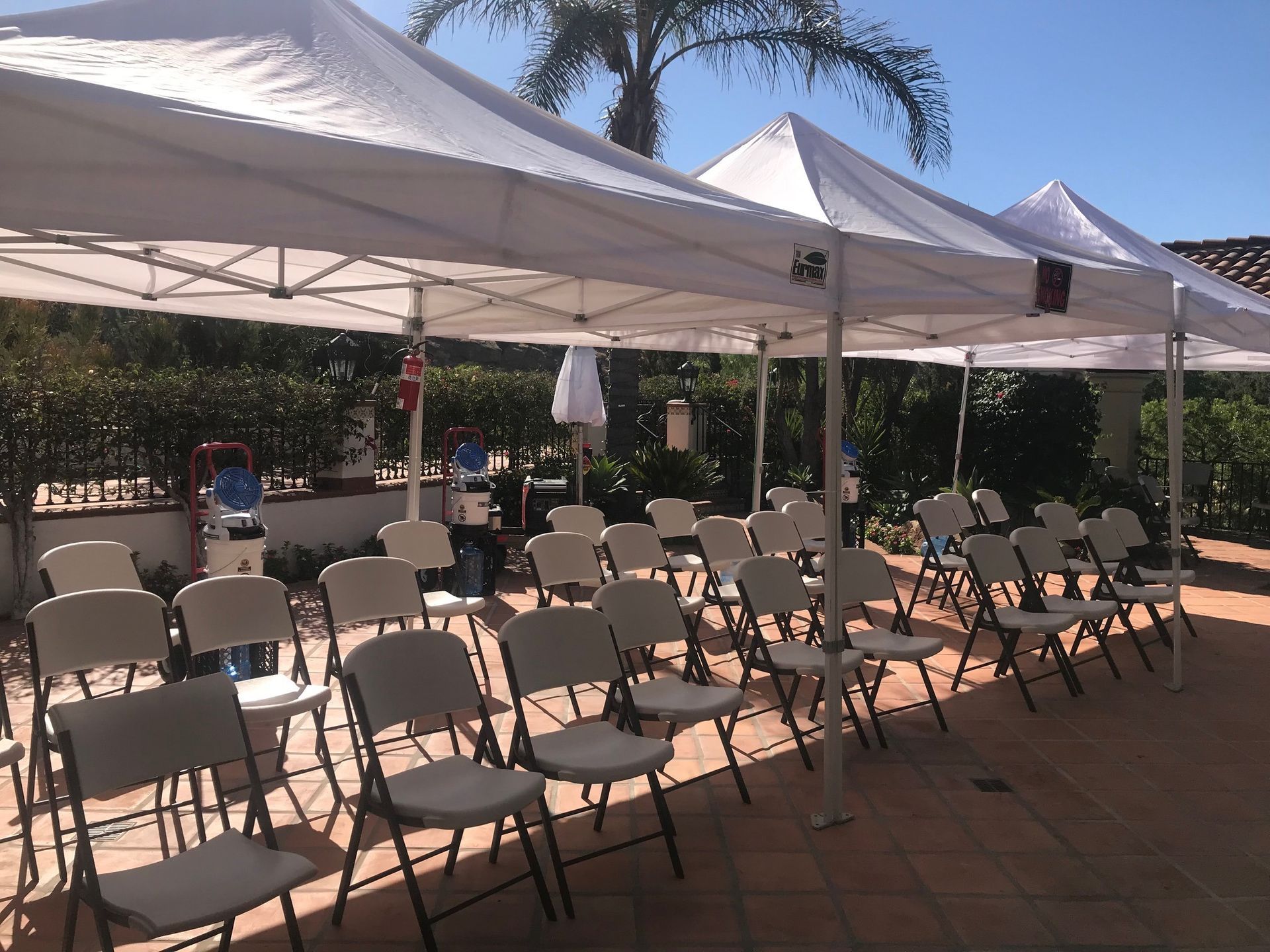 White tents sheltering rows of white folding chairs set up on a patio, likely for an outdoor event.