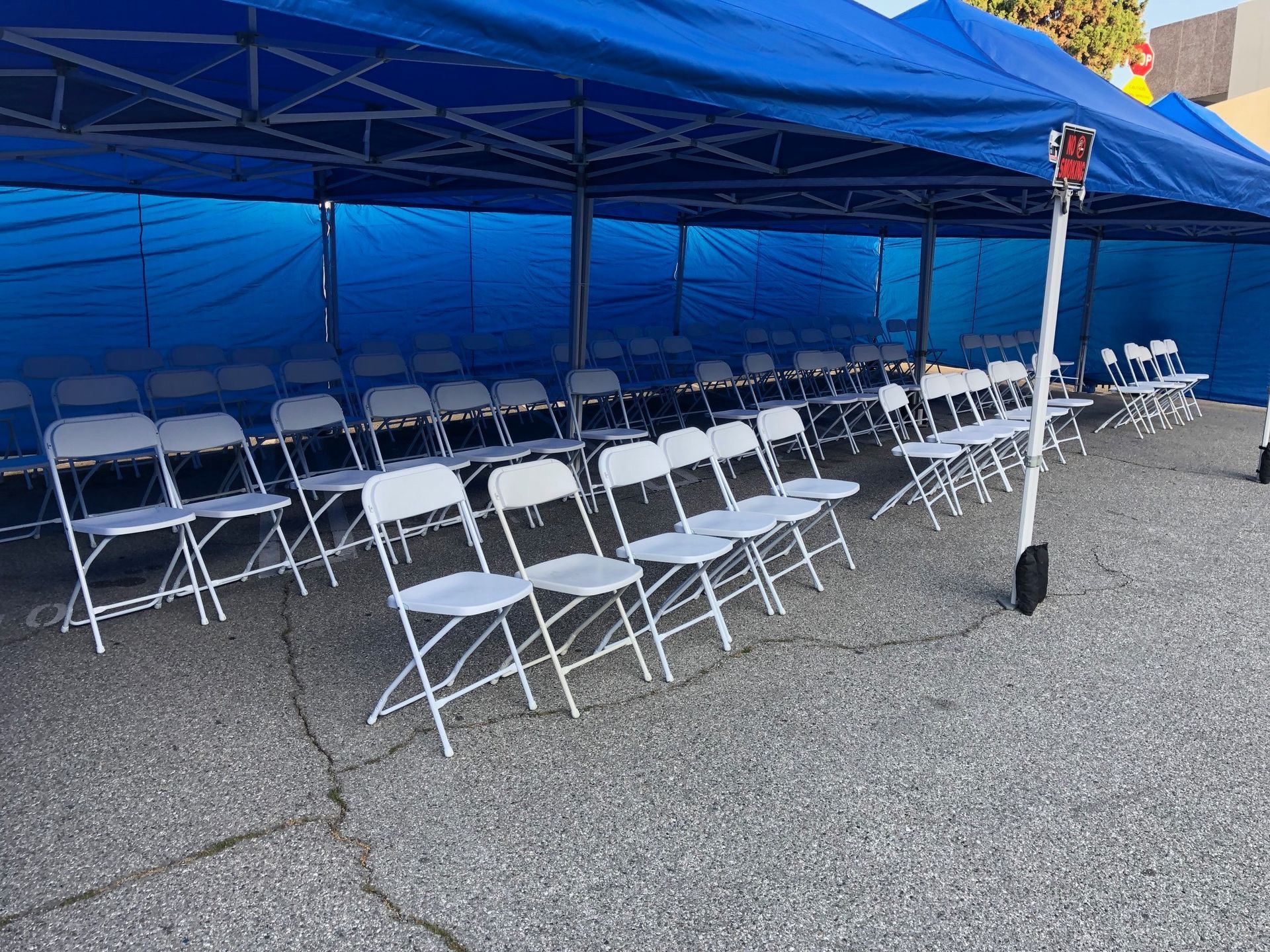Rows of white folding chairs under a blue canopy on an asphalt surface.