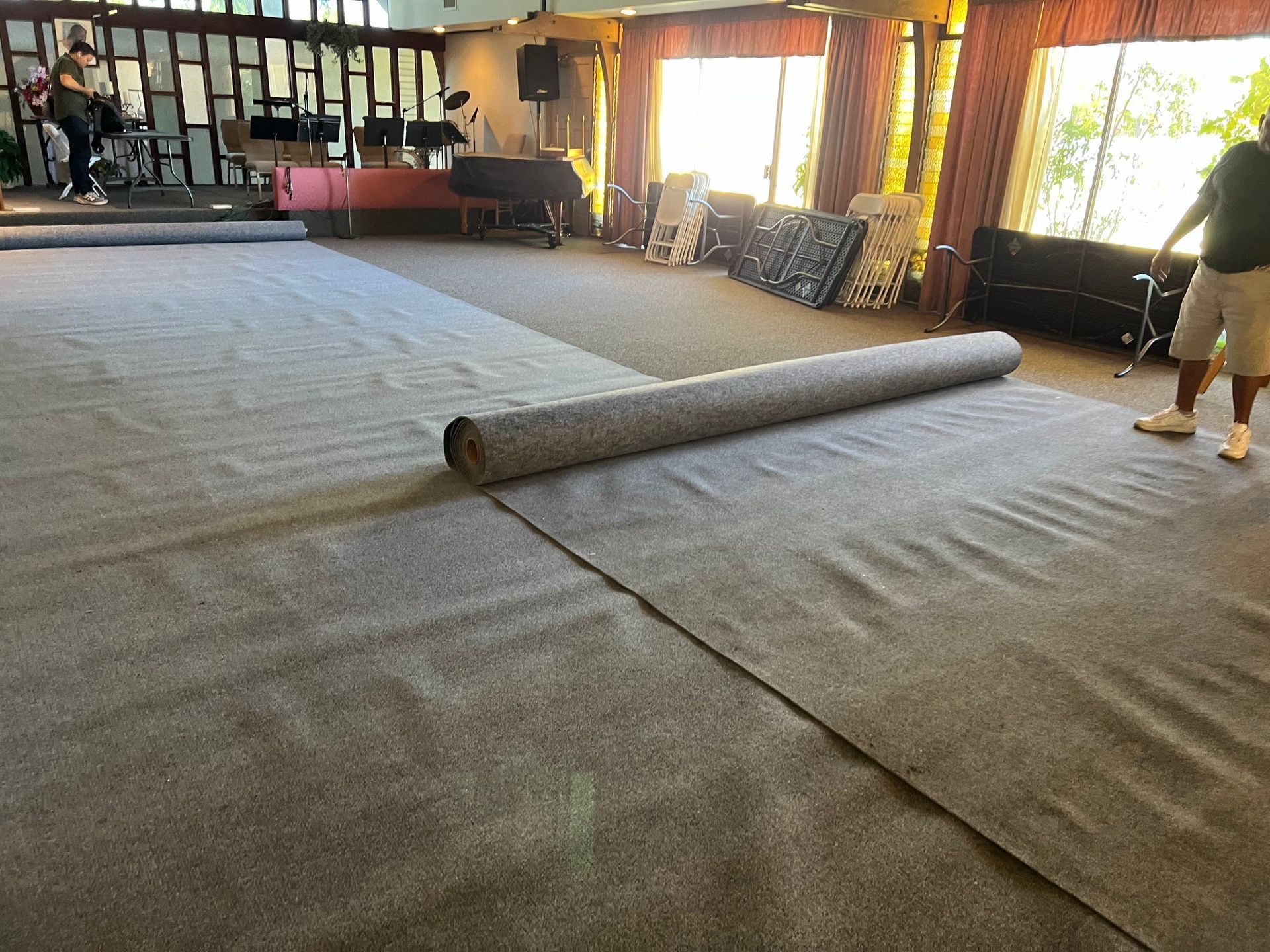 Person rolling out gray carpet in a large room, near a stage and windows.