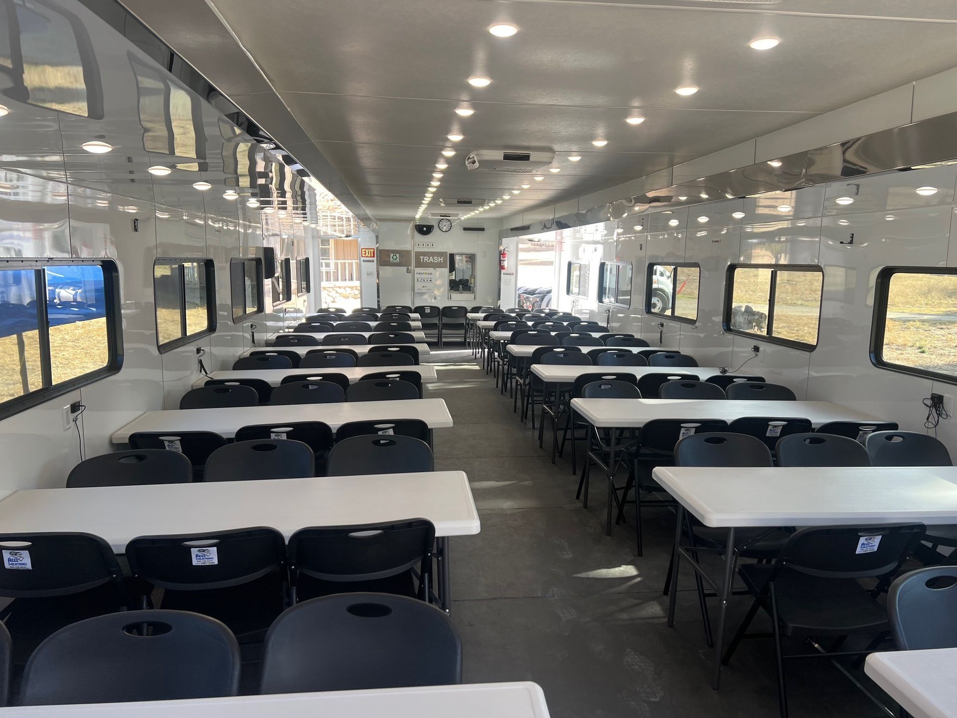 Interior view of a trailer with rows of white tables and black chairs, possibly for a meeting or event.
