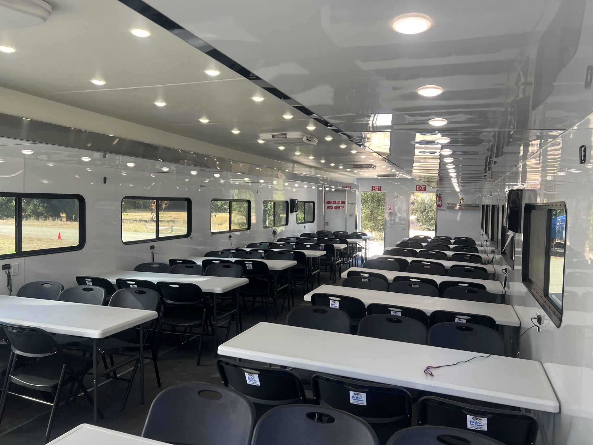 Interior of a mobile office with rows of tables, black chairs, windows, and bright lighting.