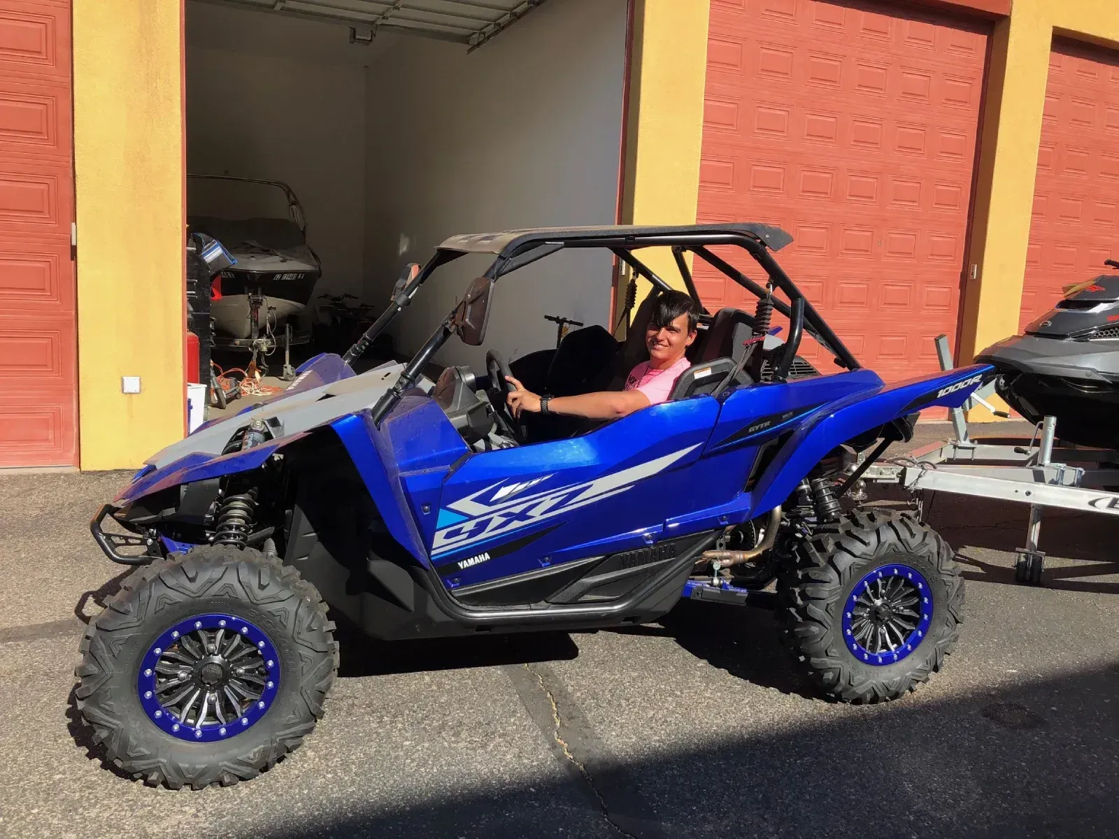 Person in blue off-road vehicle with roll cage, smiling. Parked outdoors near building with jet ski.