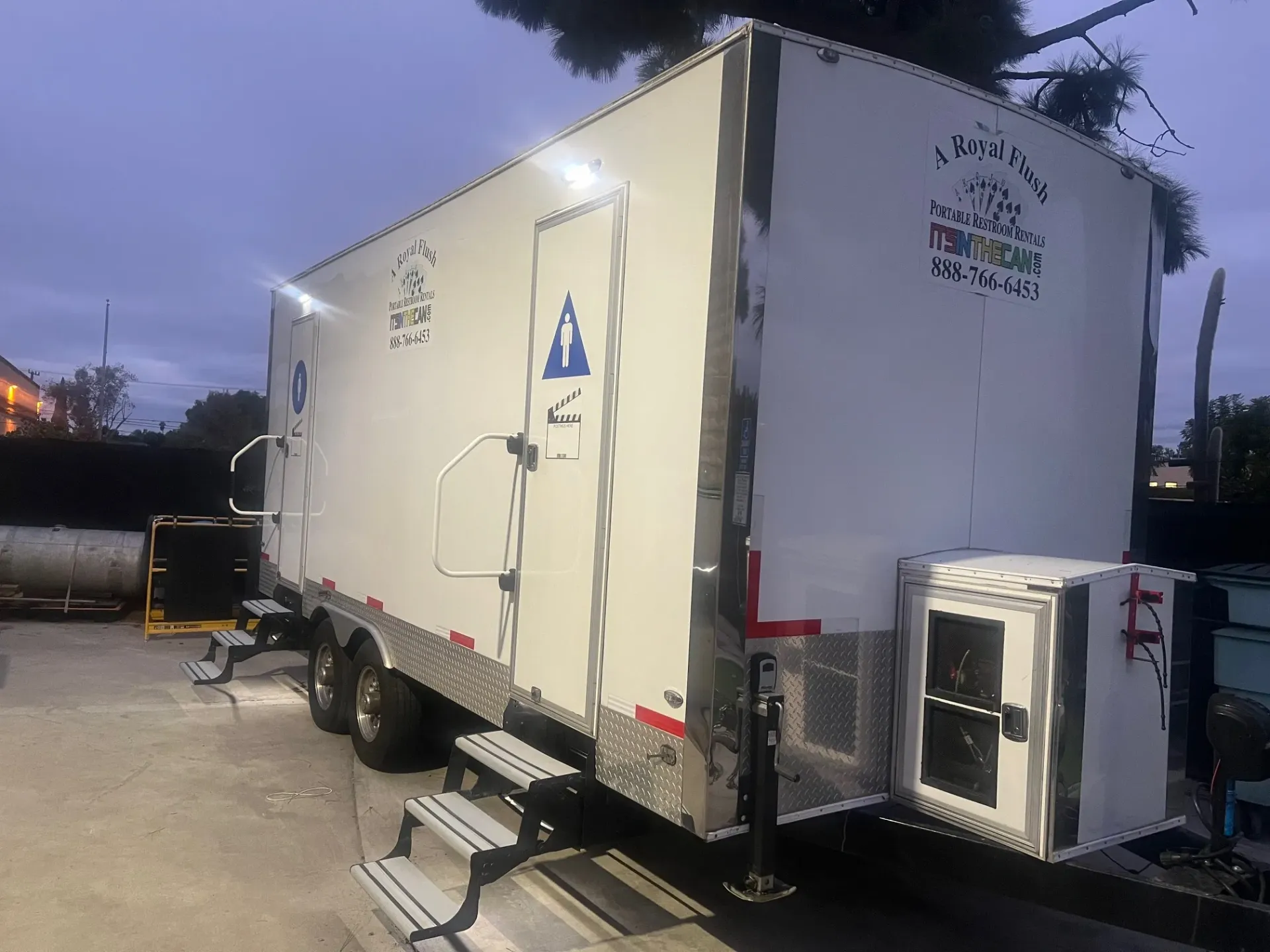 A white portable restroom trailer with steps. Located outdoors, near trees.