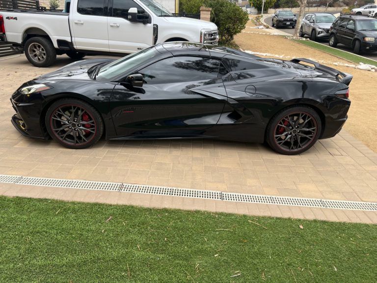 Black sports car parked on a paved driveway next to green grass, with a white pickup truck in the background.
