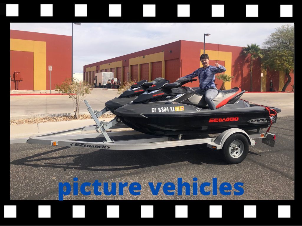 Man on two black jet skis on a trailer in front of a building; daytime.