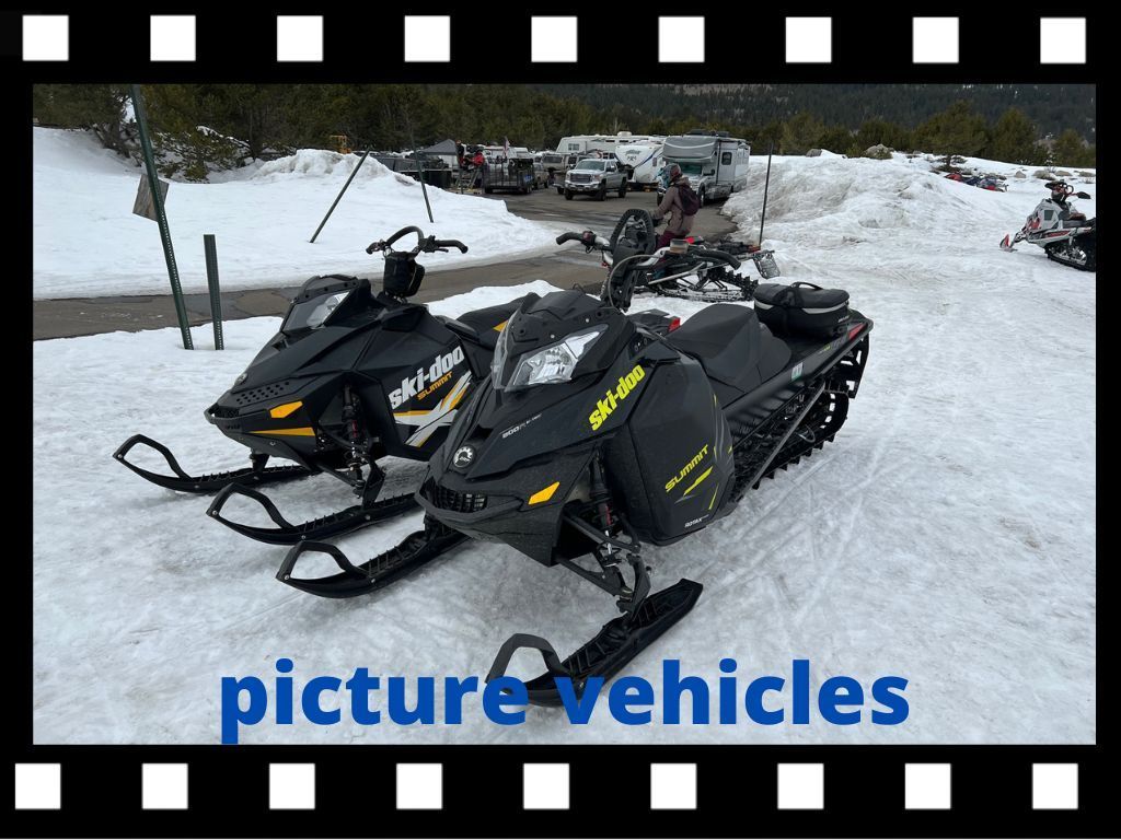 Two black snowmobiles parked on snow, other vehicles in background.