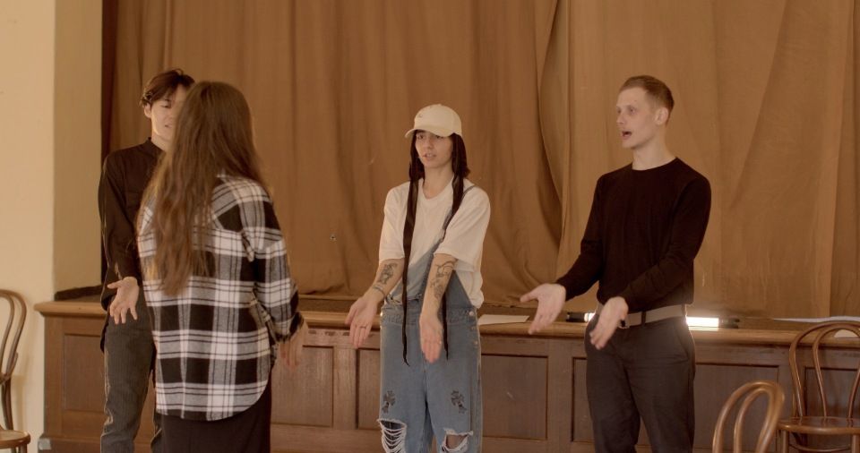 Five people on a stage, gesturing. A woman with a hat and overalls stands in the center.