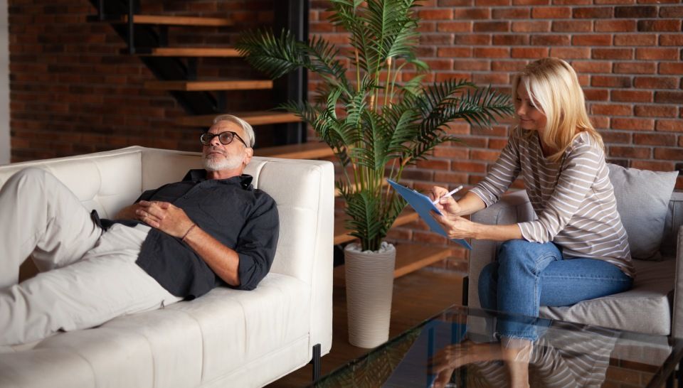 A therapist and a patient in a consultation. The man is reclining on a couch, and the woman is taking notes.