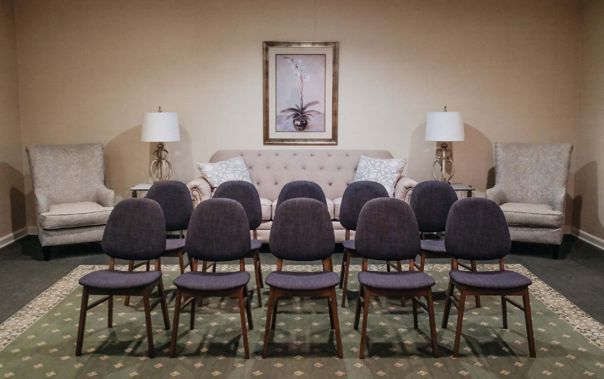 A waiting room with chairs and a sofa. Beige walls, green rug, and two lamps flank a floral painting.