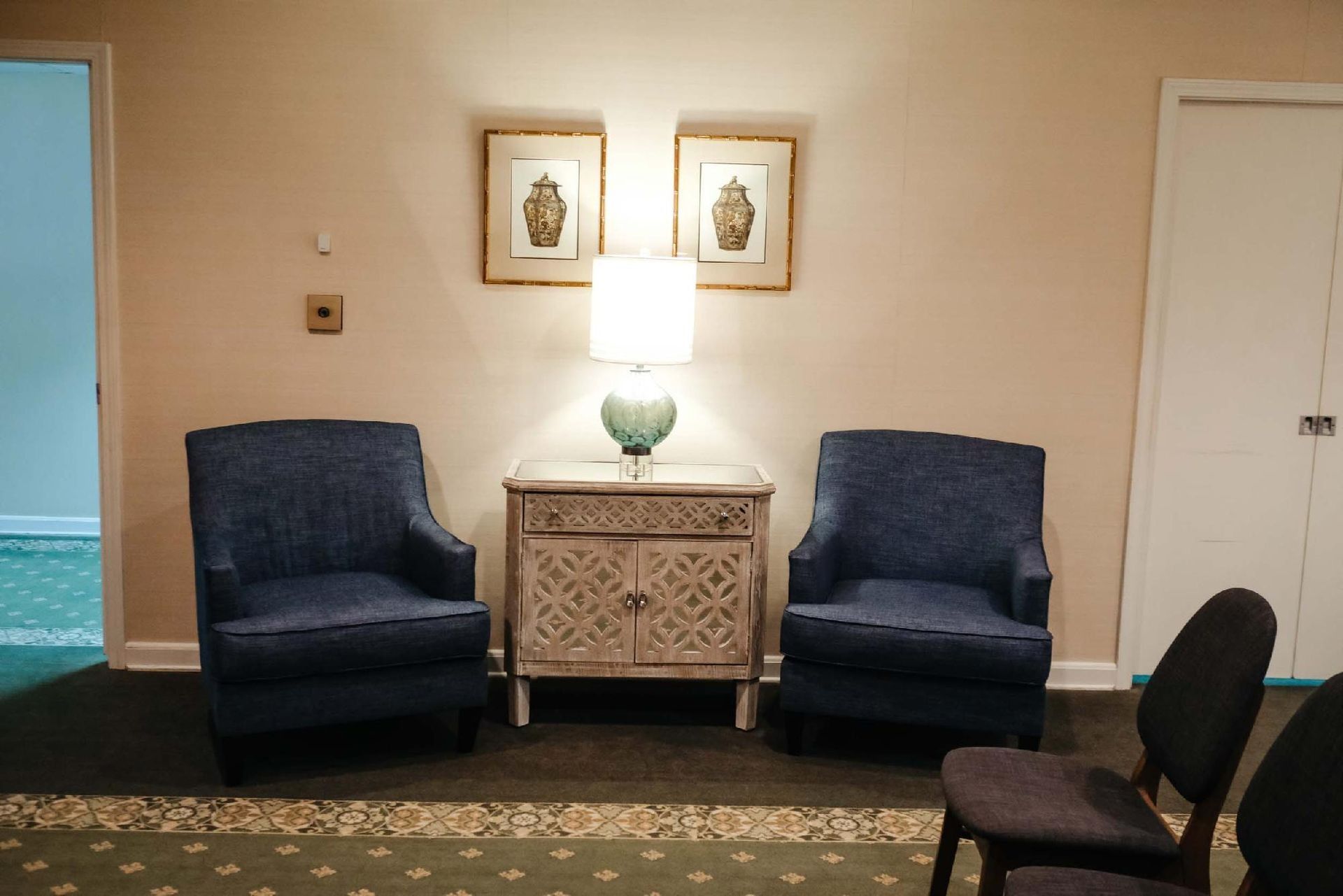 Two blue armchairs flank a decorative cabinet with a lamp, two framed prints above. A door is to the right.