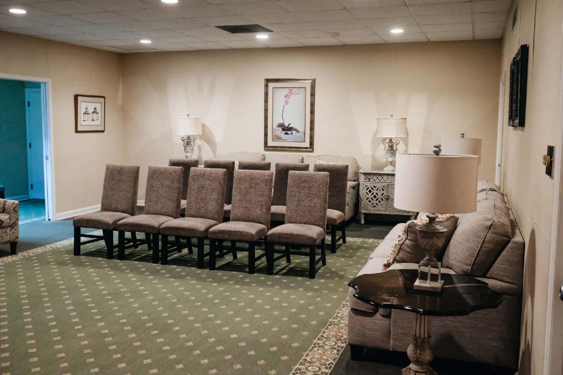 Funeral home visitation room with chairs, sofas, lamps, artwork, and green carpet.