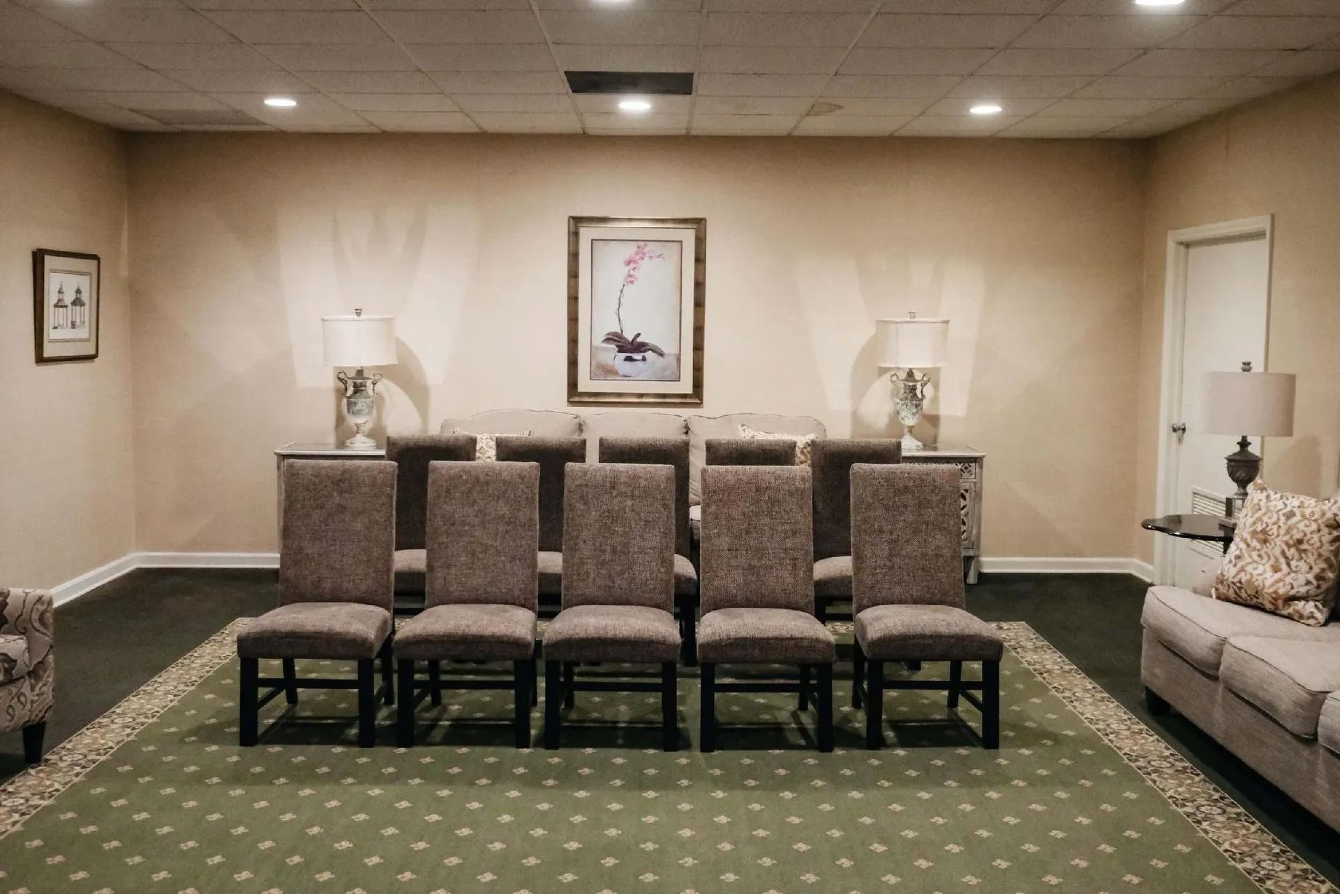 A beige room with chairs arranged facing a wall with a framed picture and two lamps.