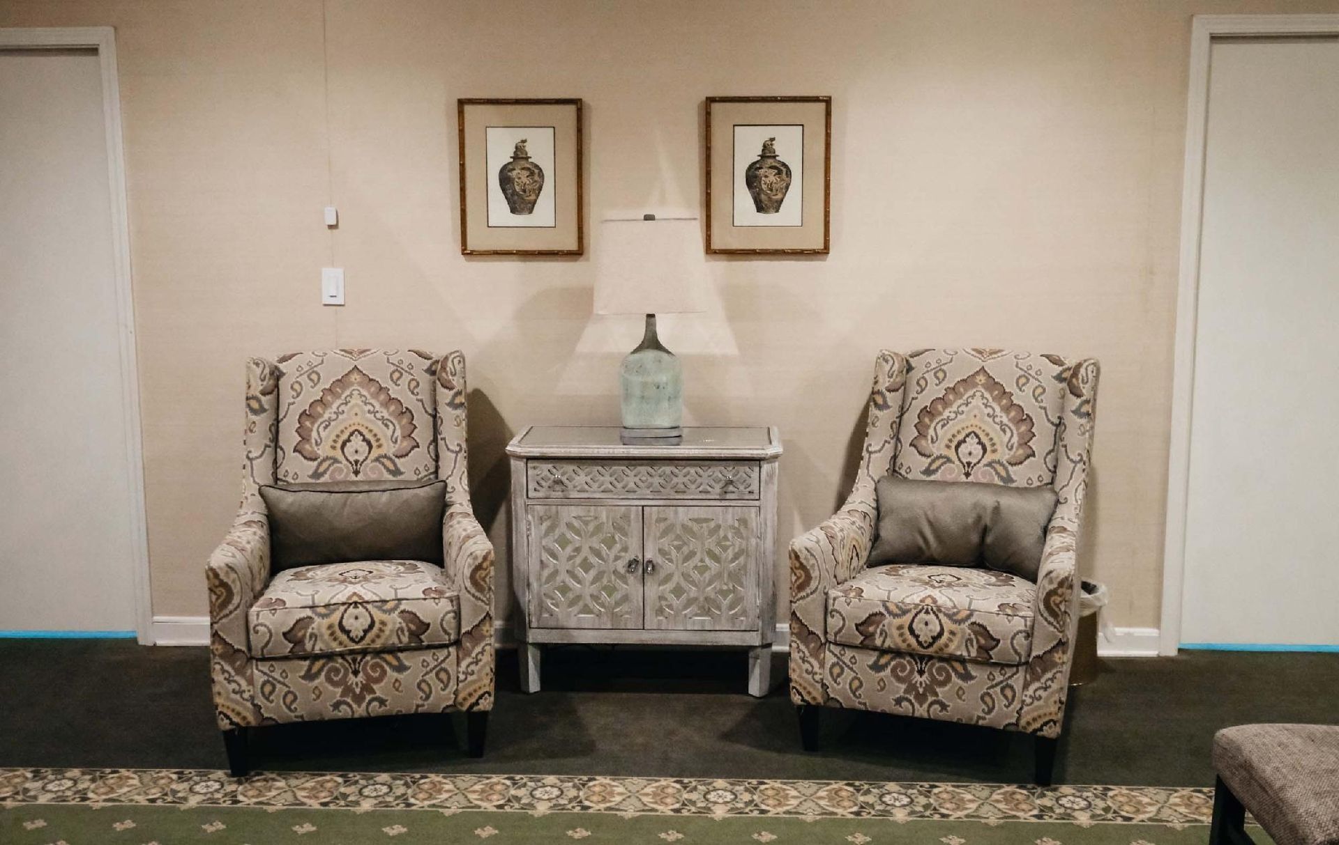 Two patterned armchairs flank a small cabinet with a lamp, artwork, and two white doors.
