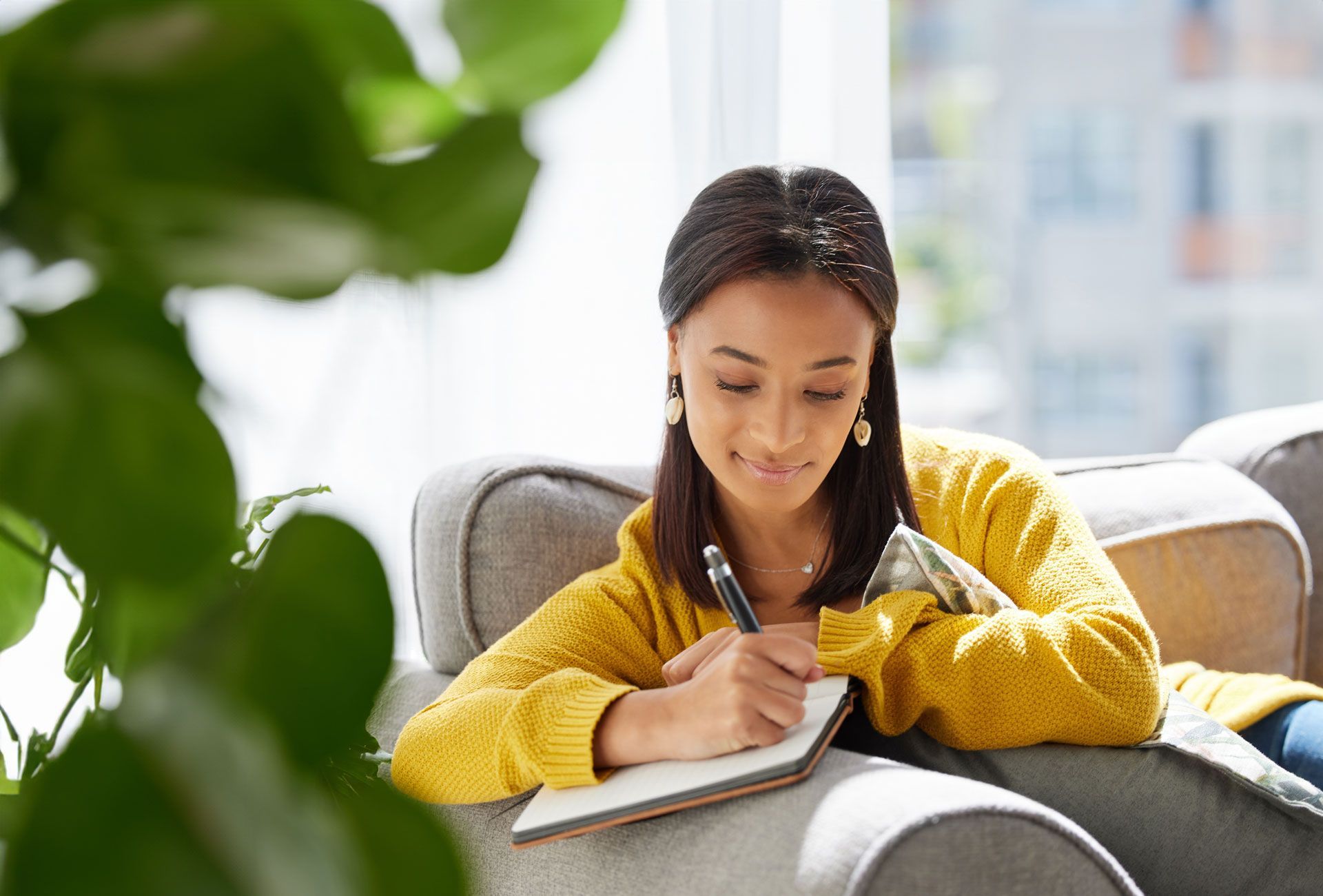 Woman in yellow sweater writes in notebook on a gray sofa, near a window, with a plant in foreground.
