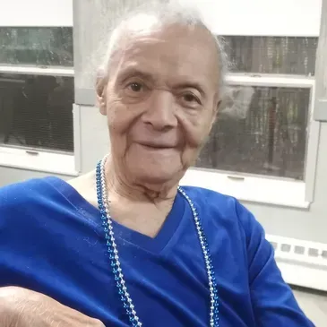 Elderly woman with fair skin wearing a blue shirt and beaded necklace smiles at the camera.